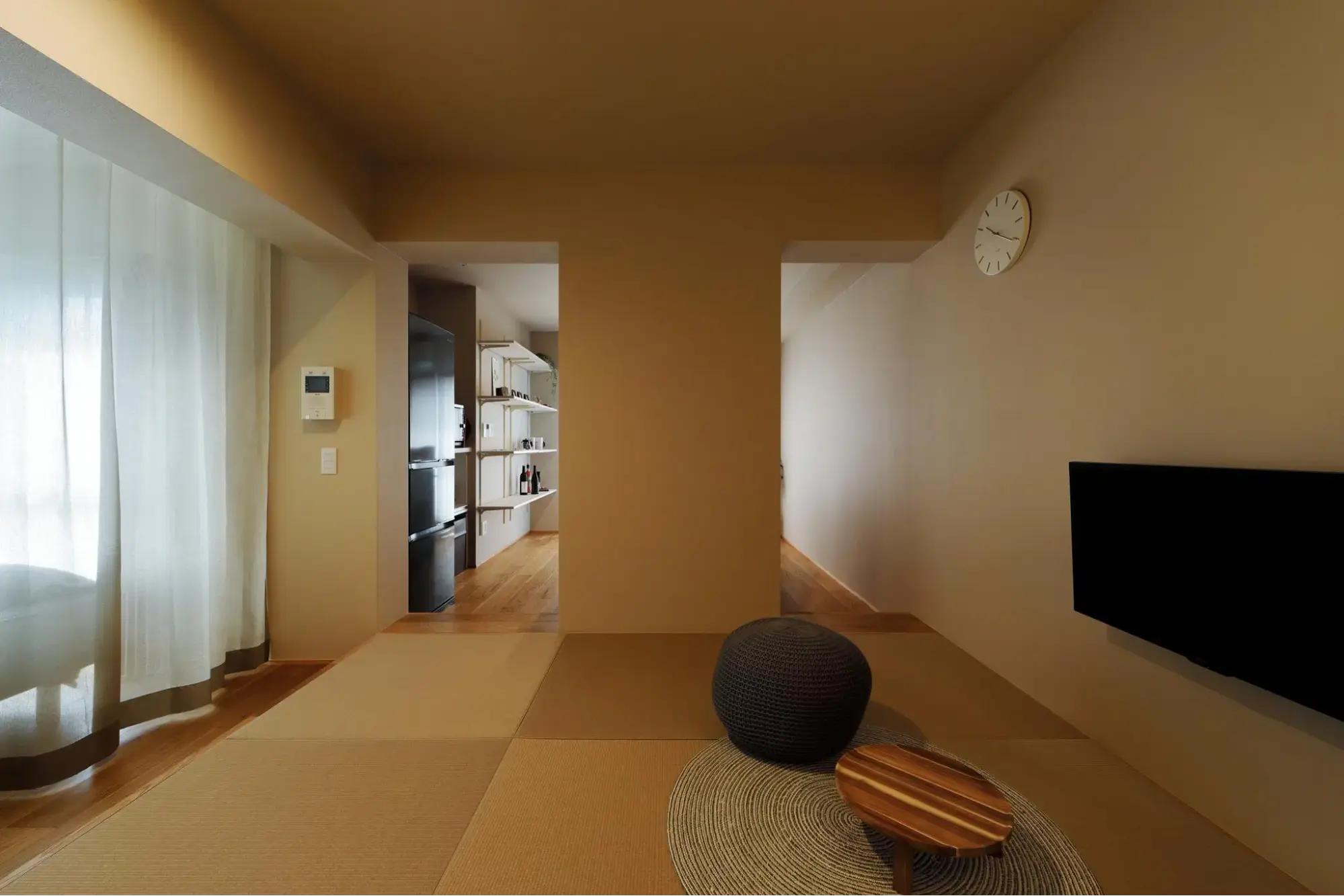 マンションフルリノベーション後。縁なし畳が敷かれたベージュ基調の空間。手前に丸いローテーブルとグレースツール、右手に壁掛けテレビを配置。奥のキッチンへ繋がる2つの開口部がある和モダンな部屋。株式会社ひかり工務店のリノベーション事例。