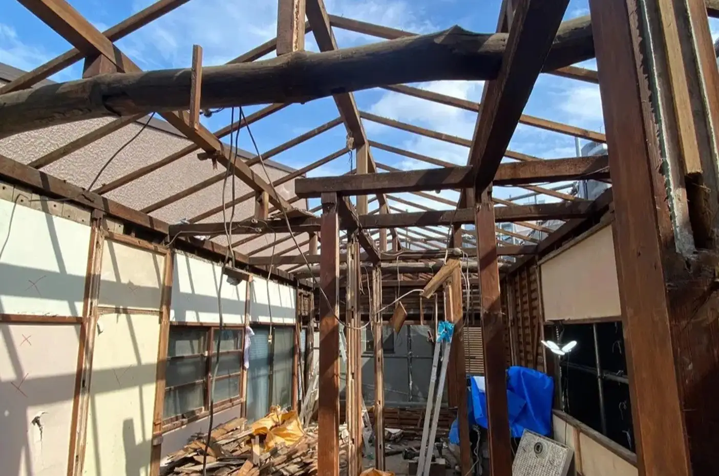 築60年古民家の解体工事中の内観写真。屋根材が撤去され、黒ずんだ既存の太い梁や束、母屋による小屋組みが完全に露出している。見上げの構図で、骨組みの隙間からは青空が覗く。耐震補強や構造再生の過程を示す記録画像