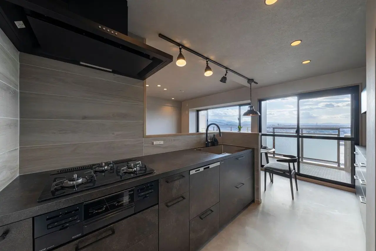 ダークグレーのグラフテクト製キッチンを中心に、黒のガスコンロと木目調の壁面タイルを配置。天井のダクトレール照明と大きな窓からの眺望が広がる、ホテルライクなモダン空間。an cubeのリノベーション事例。