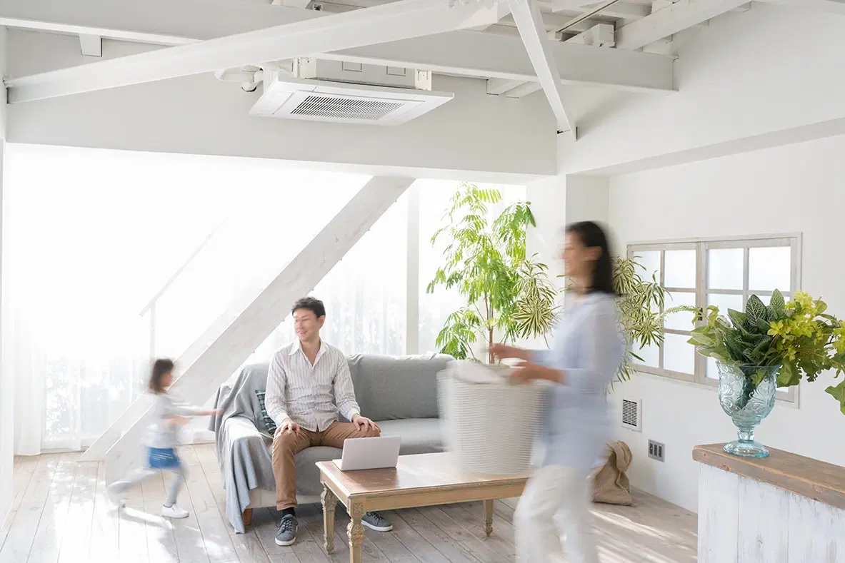 白く塗装された現し梁と高い天井が特徴的なリビング。中央にはグレーのソファと木製ローテーブルを配置し、家族がくつろぐ様子が写っている。大きな窓から自然光が入り、白い床材と観葉植物が明るい空間を構成する、リフォーム川越のイメージ画像。