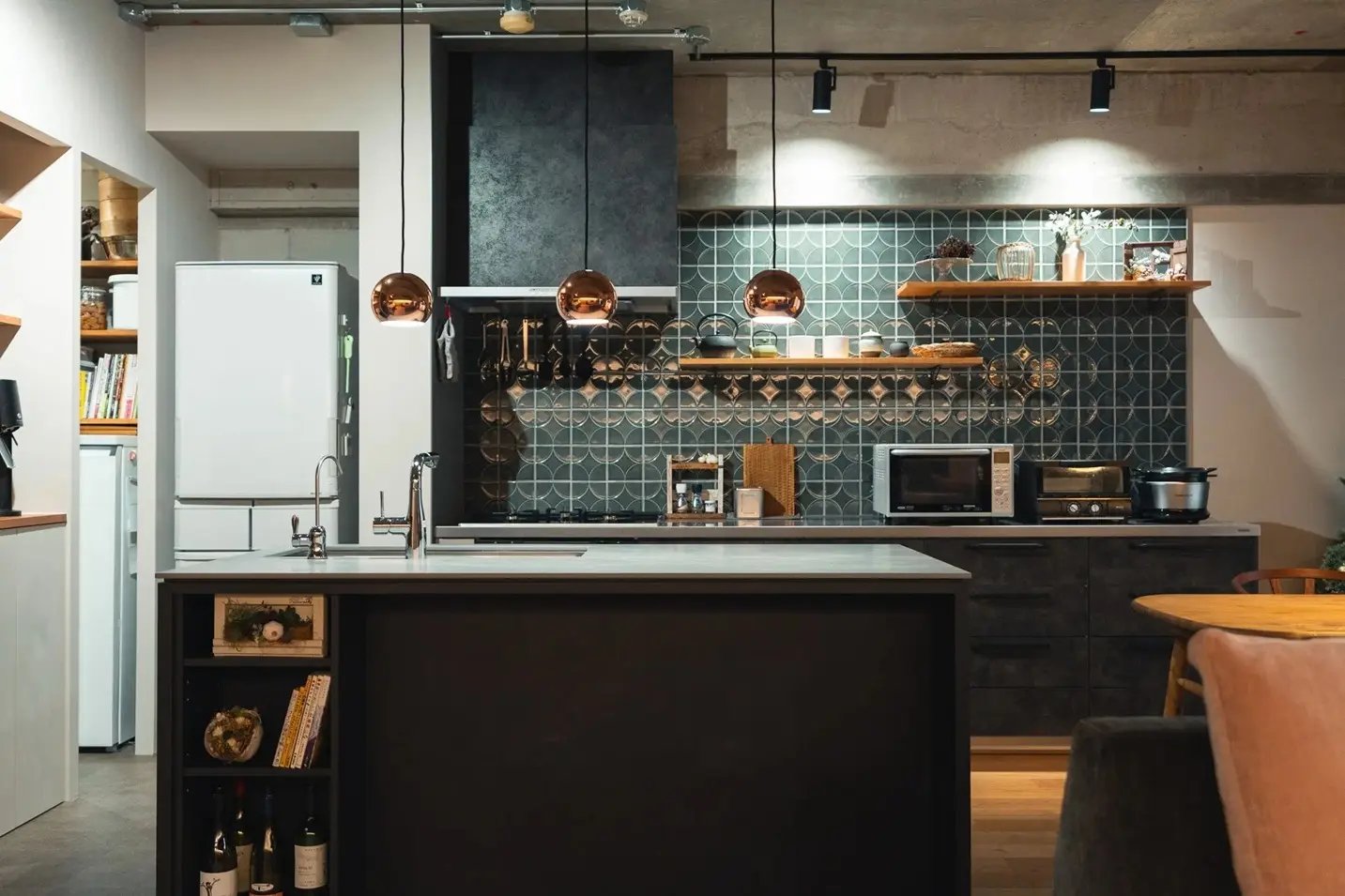 コンクリート現しの天井と、壁面の幾何学模様のタイルが特徴的なキッチン空間。黒いセラミック天板の造作アイランドキッチン上部にはカッパー色のペンダントライトが並び、背面には木製のオープン棚を配置している。可美建築の施工事例。