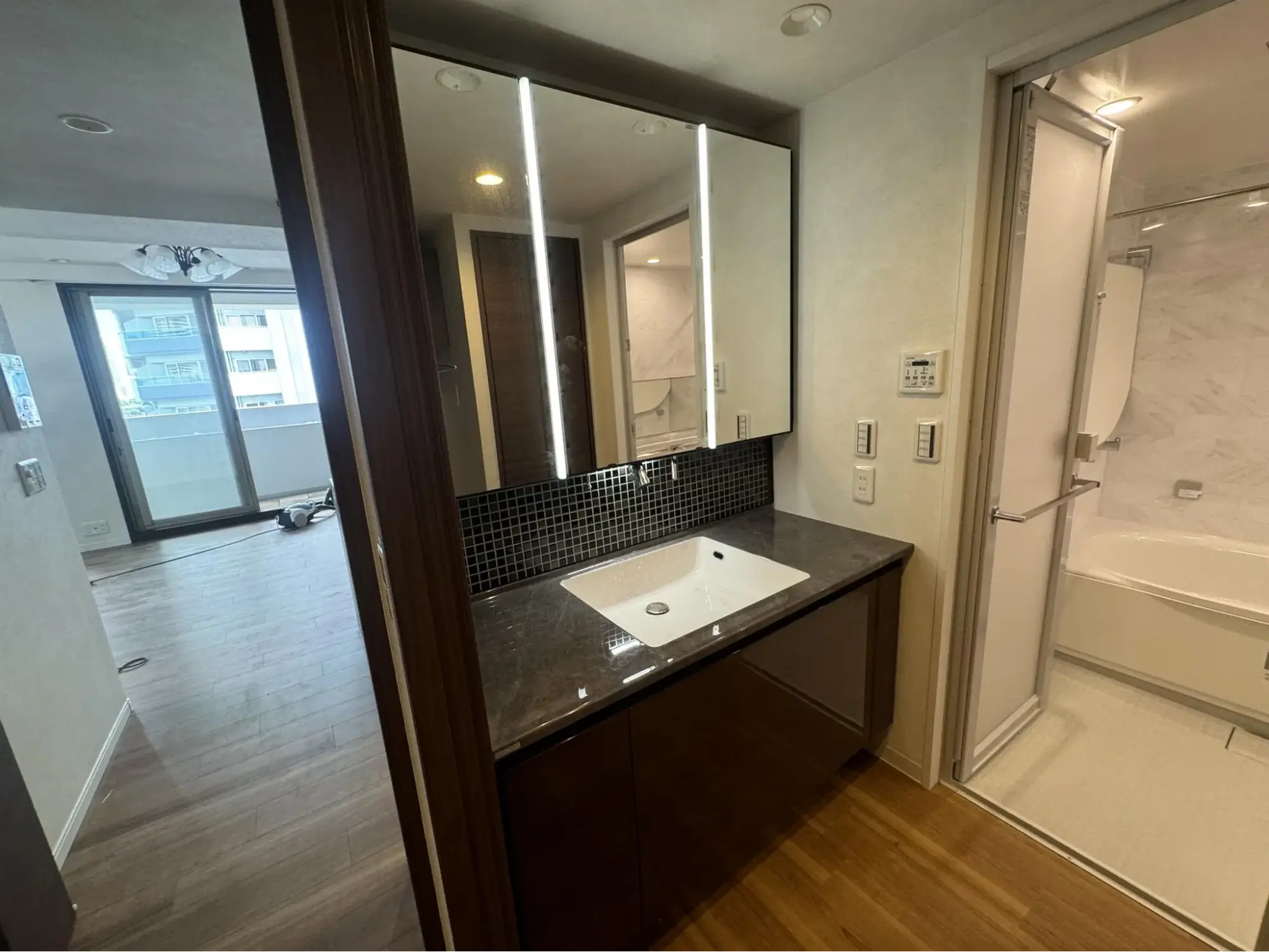 縦型の照明が内蔵されたスタイリッシュな三面鏡と、ダークカラーの天板や黒のモザイクタイルを組み合わせた洗練された洗面台。水回り設備にこだわり、程よい大人っぽさとホテルライクな高級感を演出した空間。リフレネクスト横浜による中古マンションのフルリノベーション事例。