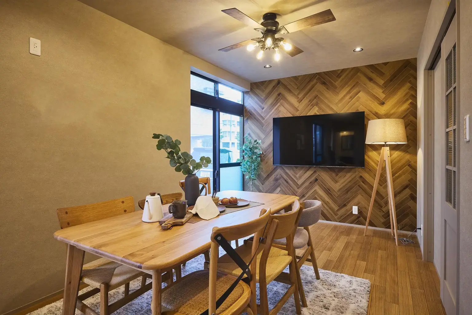 V字の木目が美しいヘリンボーンのアクセントウォールが目を引くダイニング。壁掛けテレビや木製家具、シーリングファンを合わせ、温かみのある空間を演出。元和室から現代風のLDKへとフルリノベーションした、My Design/株式会社Izumidaの八王子スタジオ