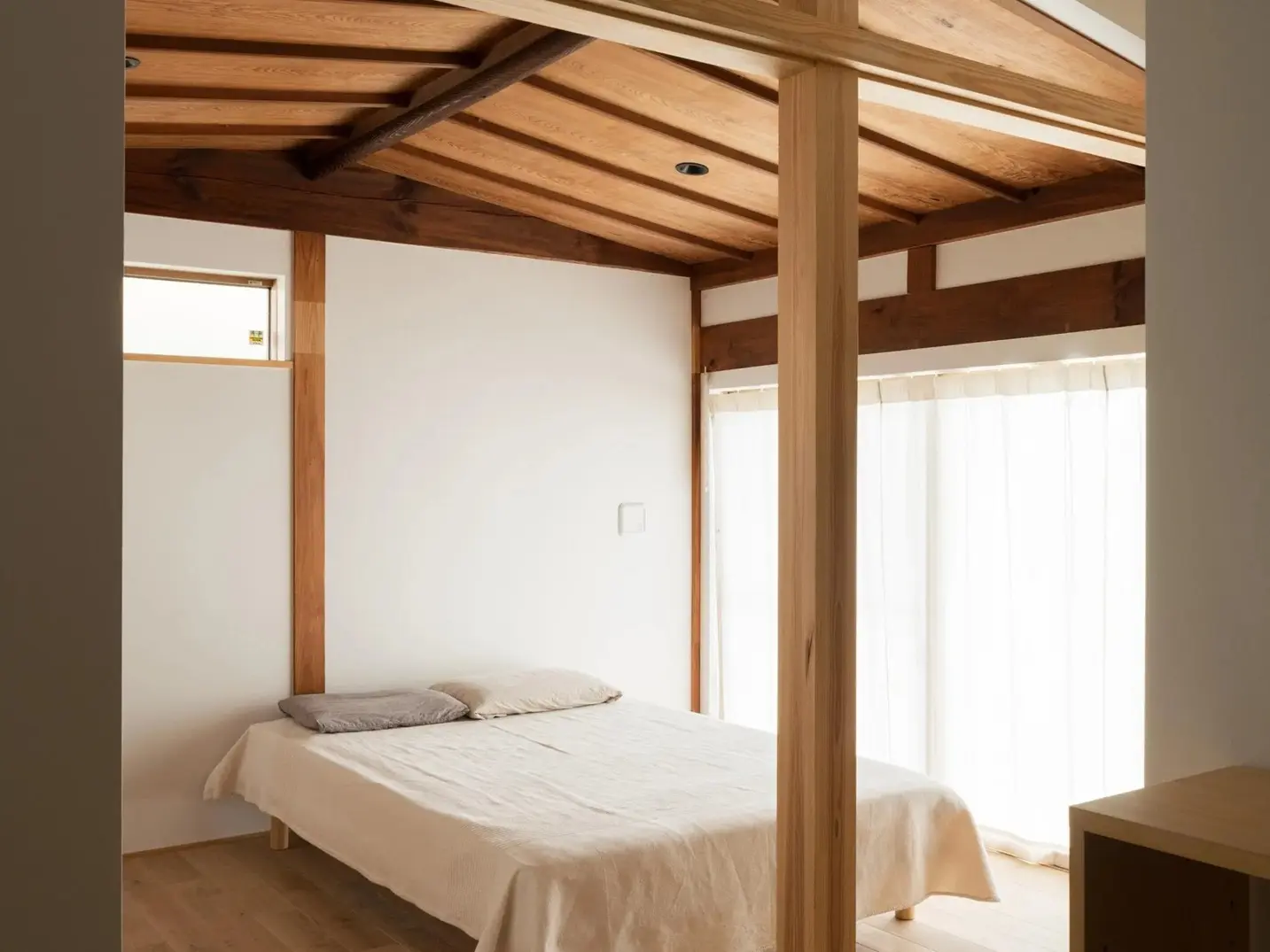 現しの垂木が美しい勾配天井と、部屋の中央に立つ太い木の柱が特徴的な寝室。白い壁に囲まれ、大きな窓から自然光が柔らかく差し込む、シンプルで温かみのある古民家リノベーション後の内観。