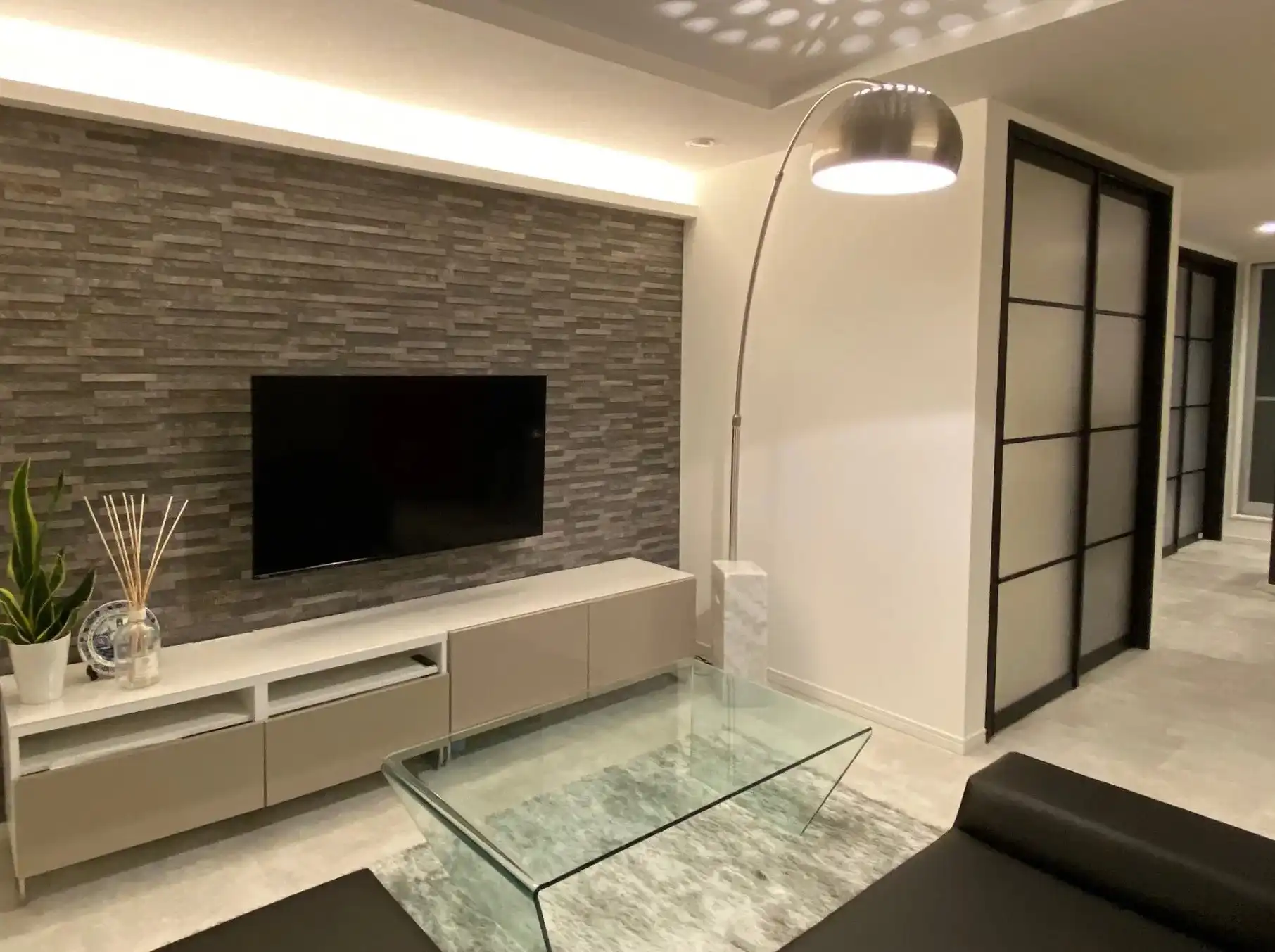 グレーのタイル(エコカラット)を貼った壁があるリビング。壁掛けテレビ、テレビ台、ガラスのコーヒーテーブルが置かれている。