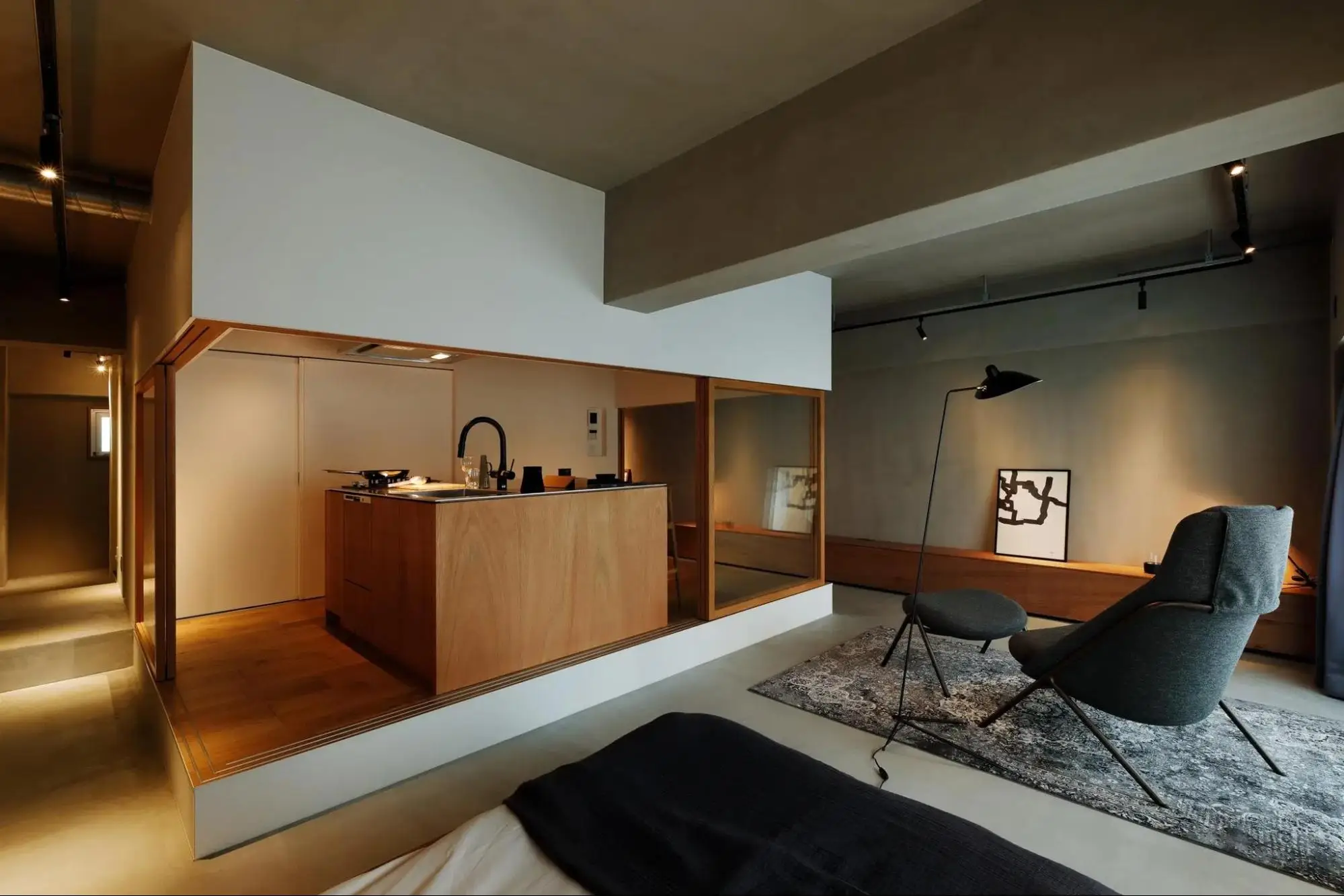 ワンルームの室内写真。中央に床より一段高い、板張りの小上がりキッチンが配置され、手前のコンクリート床にはベッドとラウンジチェアが置かれている。