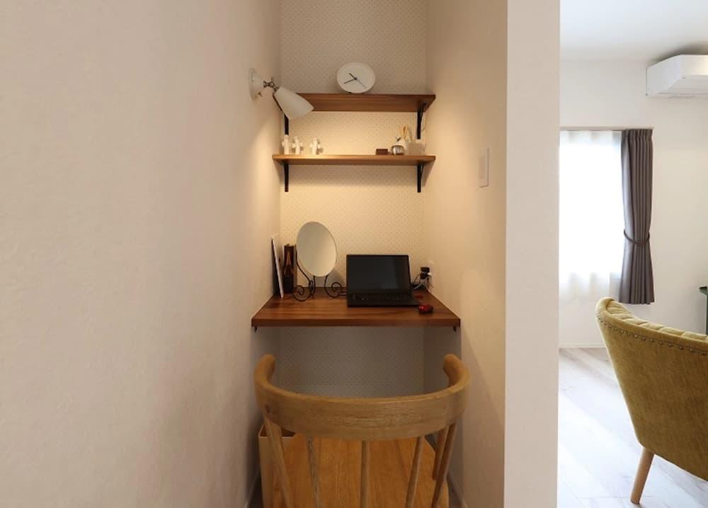 白い壁に囲まれた小さなくぼみを活用し、デスクと収納を完備したミニマルなワークスペース。落ち着いた色味の木製天板が壁に固定され、その上にはノートパソコンや丸いスタンドミラー、小物が整然と並んでいる。正面の壁にはドット柄のような控えめなアクセントクロスが貼られ、黒いブラケットで支えられた2段の木製シェルフには、白い置き時計やインテリア雑貨が飾られている。左側の壁には白い首振り式のスポットライトが設置され、手元を明るく照らす。手前には丸みを帯びた背もたれが特徴的な木製チェアが置かれ、右側に見えるリビングの明るい日差しやイエローのソファと心地よい距離感を保っている空間。