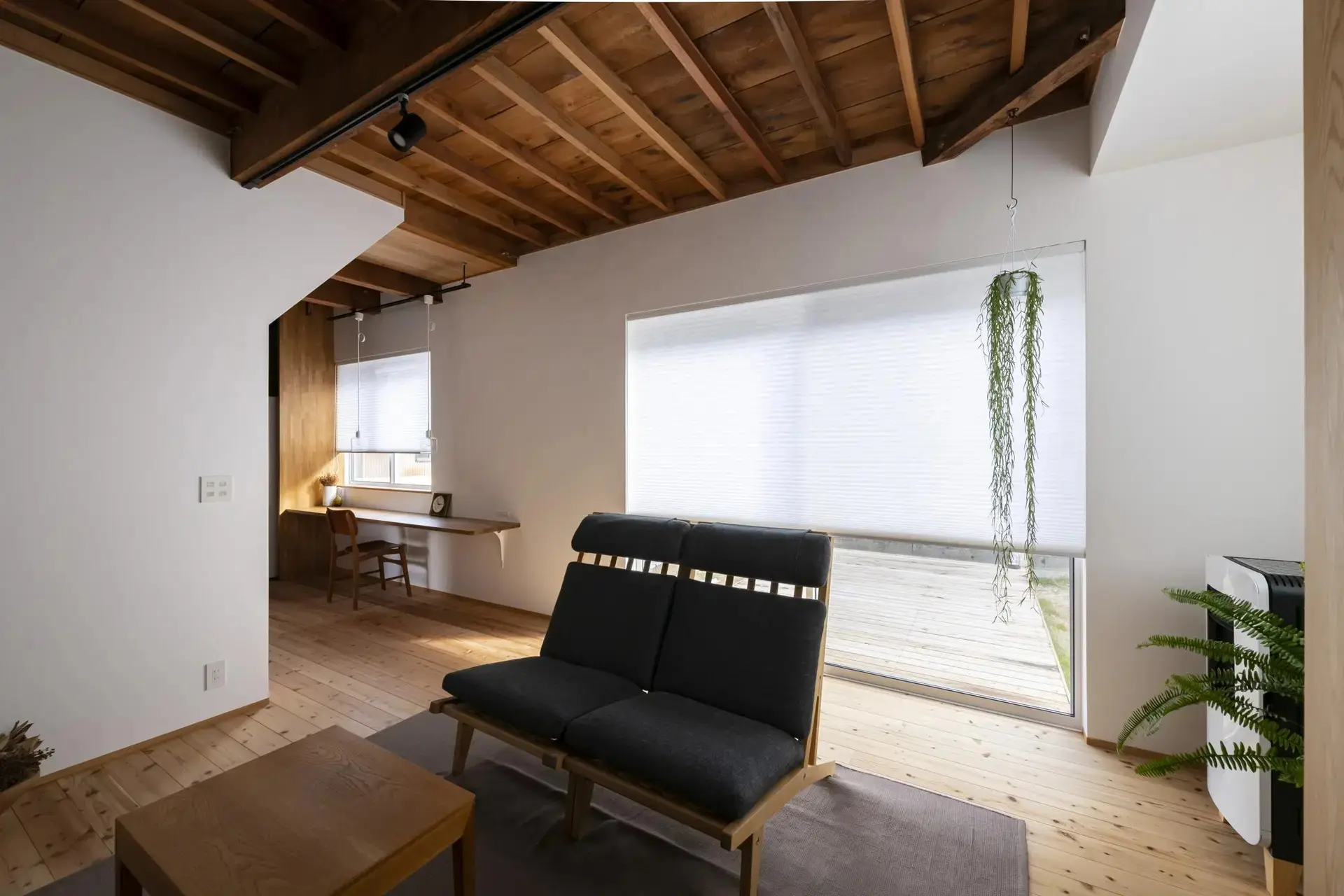 リノベーション後のリビングルーム。天井は木製の板張りで、床も木製フローリング。奥に大きな掃き出し窓があり、手前にグレーのソファが置かれている。