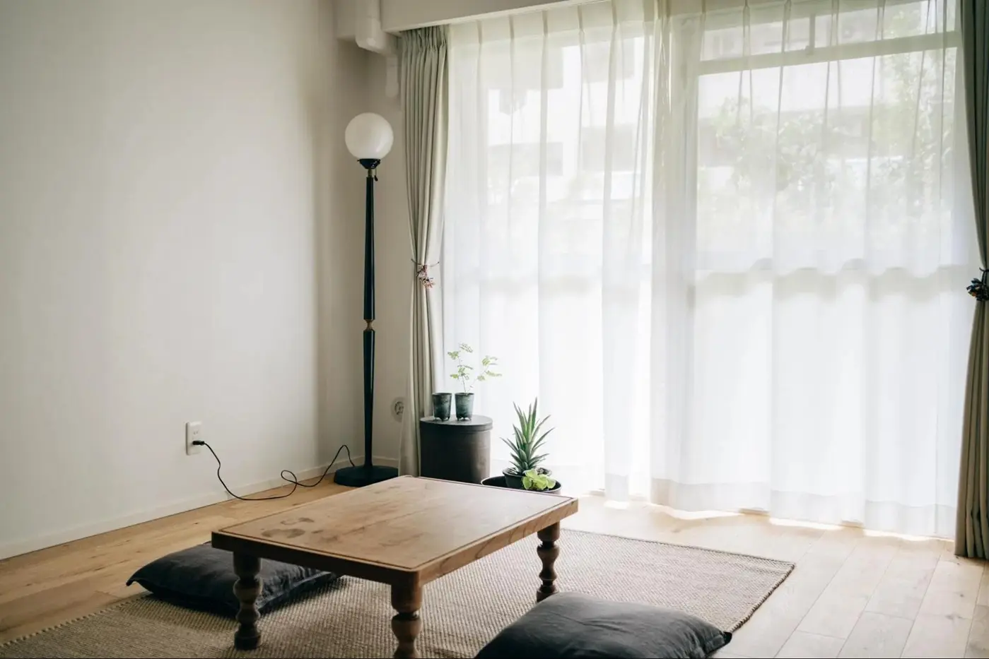 自然光が柔らかく差し込むシンプルなリビングの一角。アンティーク調の木製ローテーブルやスタンドライトが配置され、無垢材のフローリングと調和している。