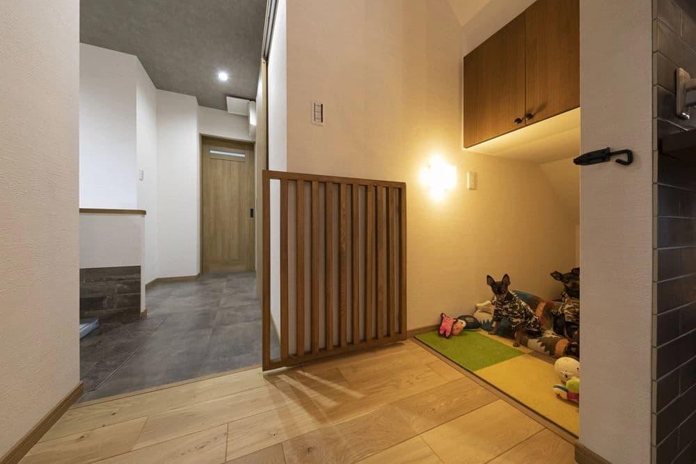 階段下の三角形の空間を有効活用した、2匹の愛犬専用のヌックスペース。入り口にはインテリアに馴染む木製の格子ゲート（ドッグゲート）が設置され、安全性とデザイン性を両立させている。内部には黄色や緑のカラフルなマットが敷かれ、カモフラージュ柄の服を着た2匹の小型犬がおもちゃに囲まれてリラックスしている様子が伺える。壁面には暖色のブラケットライト、上部の造作収納の下には間接照明が備え付けられ、夜間でも愛犬が落ち着いて過ごせる隠れ家のような演出がなされている。手前の明るい木目調のフローリングから奥のモダンなダークグレーのタイルへと切り替わるスタイリッシュな空間の中に、ペットへの愛情が詰まった設計。