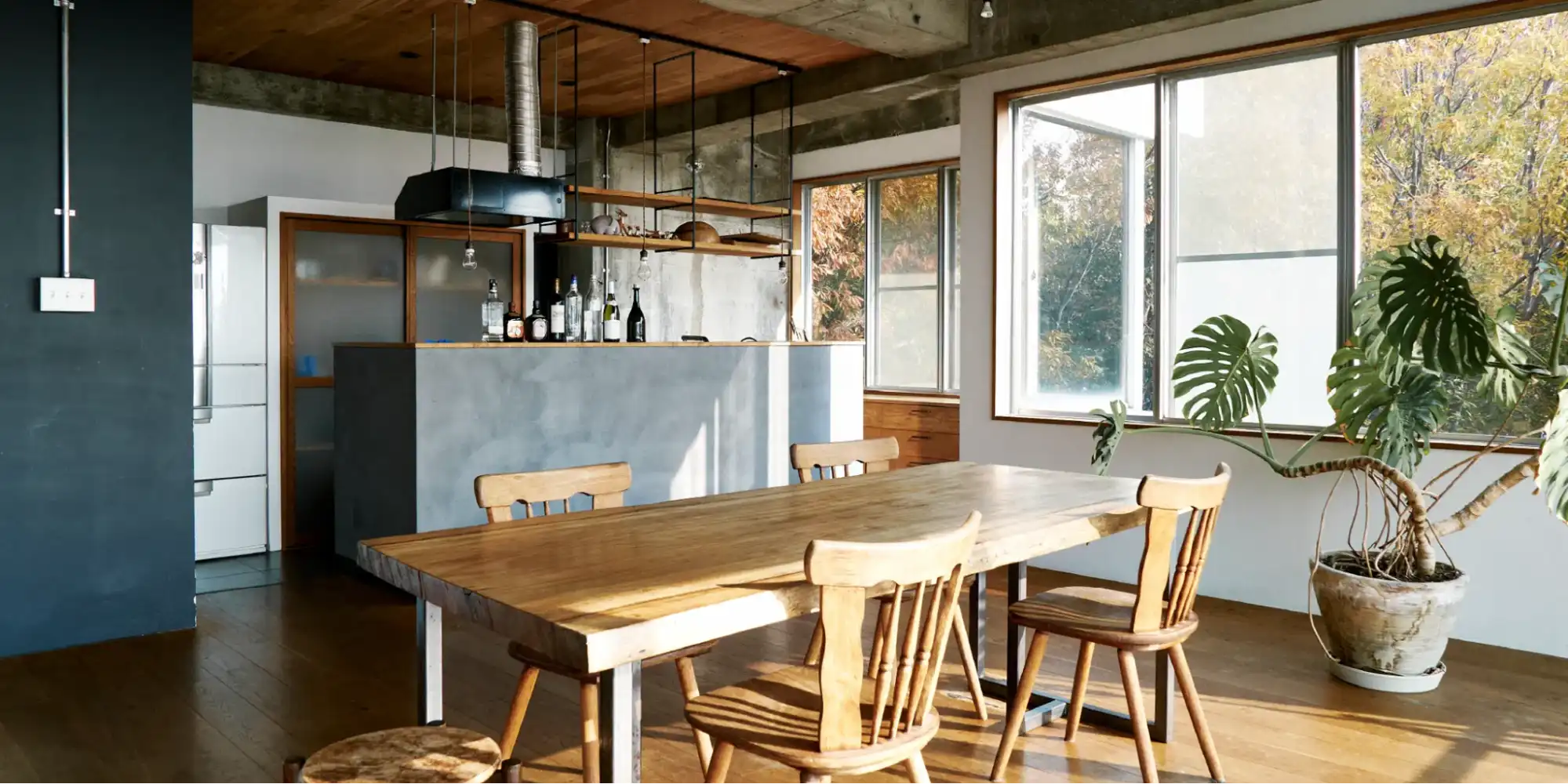 コンクリート壁と一部木張り天井のLDKリノベーション後。手前に無垢材ダイニングテーブルと木製チェア、奥にグレーの対面キッチンを配置。窓からの自然と観葉植物が調和する無骨な空間。株式会社スクエアのリノベーション事例。