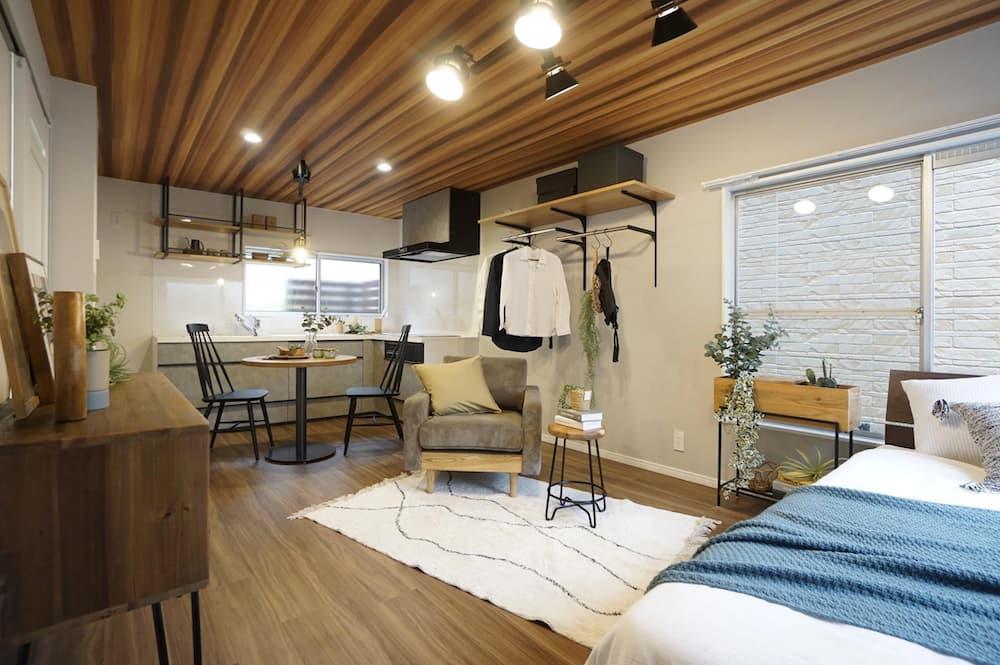 木目調の天井に黒いダクトレールとスポットライトが設置されたアパートのLDK。白いシステムキッチンの背面にはダークグレーのクロスを張り、黒いフレームの室内窓が空間のアクセントになっている。単身男性向けのインダストリアルな内装デザイン。