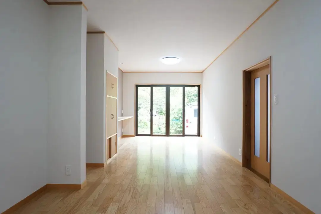 部屋の奥に設置された大きな掃き出し窓と、そこから見える庭の木々。手前にはリノベーションで新しくなった明るい木目のフローリングが広がっている。