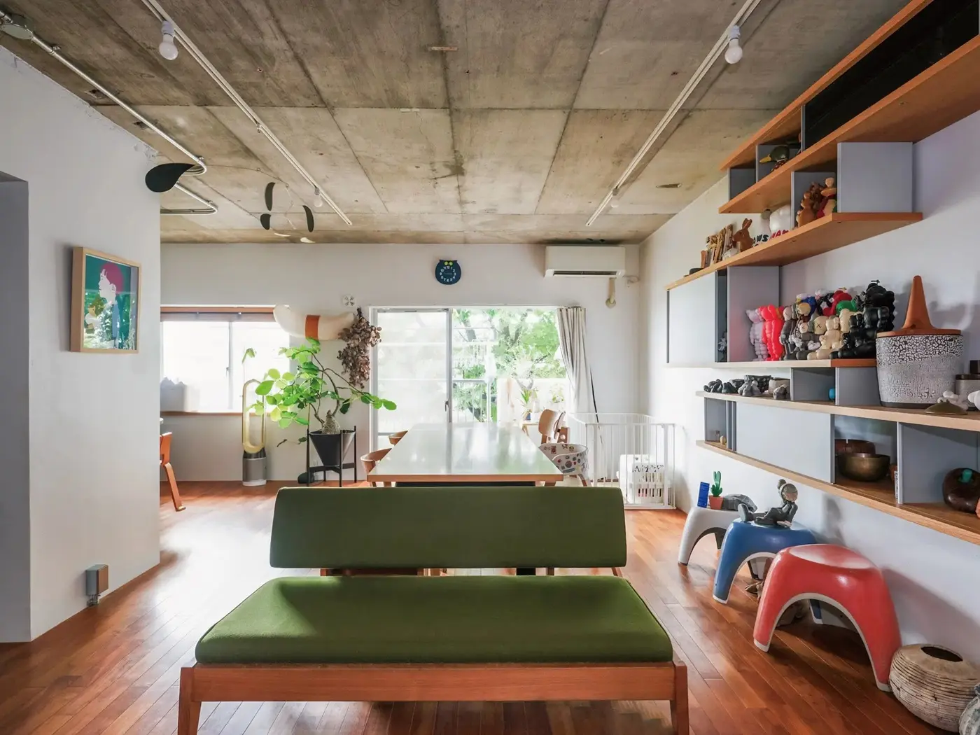 コンクリート現しの天井とフローリングで構成されたLDK。壁面の可動棚には多数のフィギュアや雑貨が並び、中央には木製のダイニングテーブルとグリーンのベンチソファが配置されている。田中建設の施工事例。