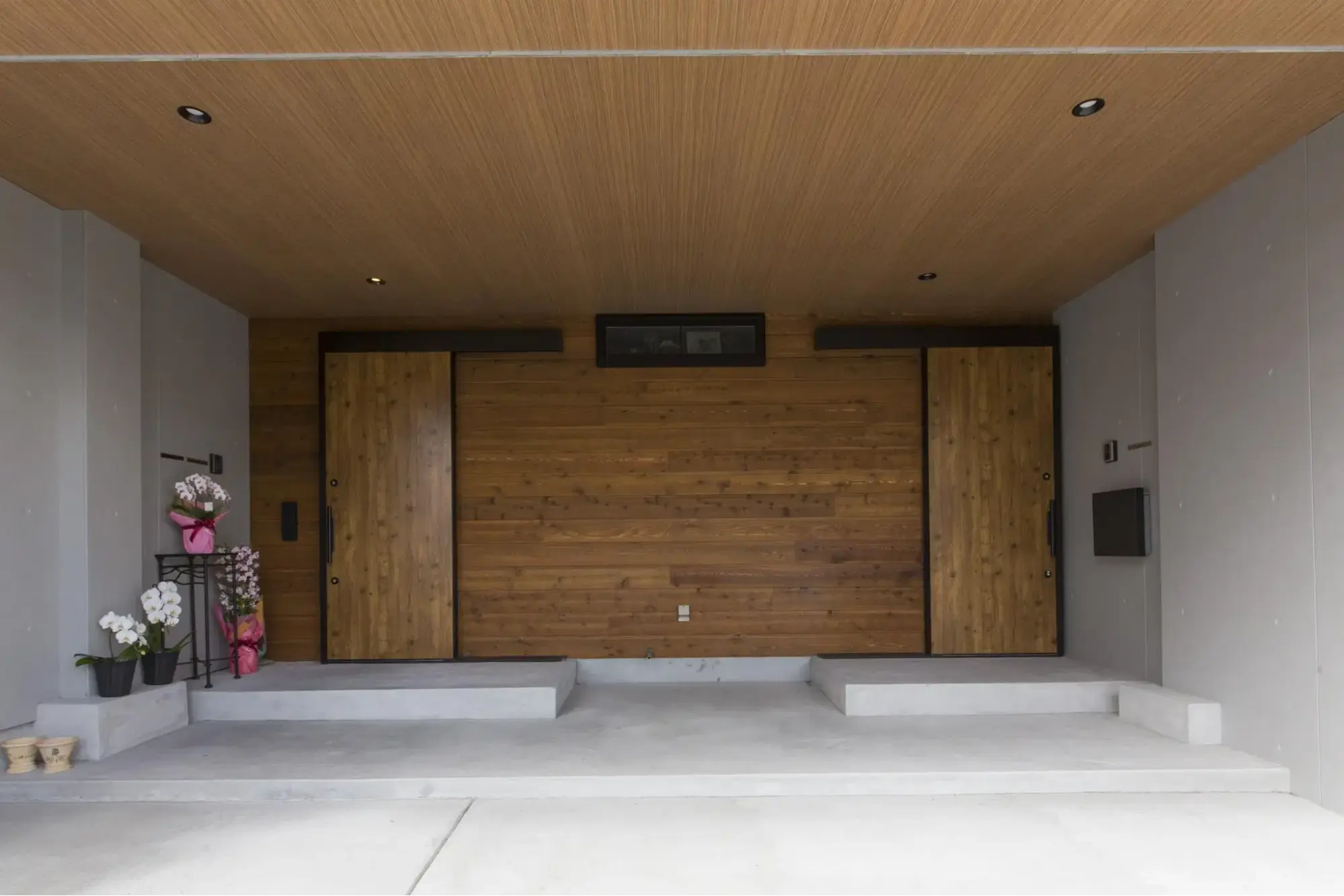 二世帯住宅のフルリノベーション後。コンクリートの壁と床が広がる深い軒下空間。木目の天井と正面の板張り外壁に、世帯ごとに独立した2つの木製引き戸が並ぶ玄関アプローチ。パパママハウスのリノベーション事例。
