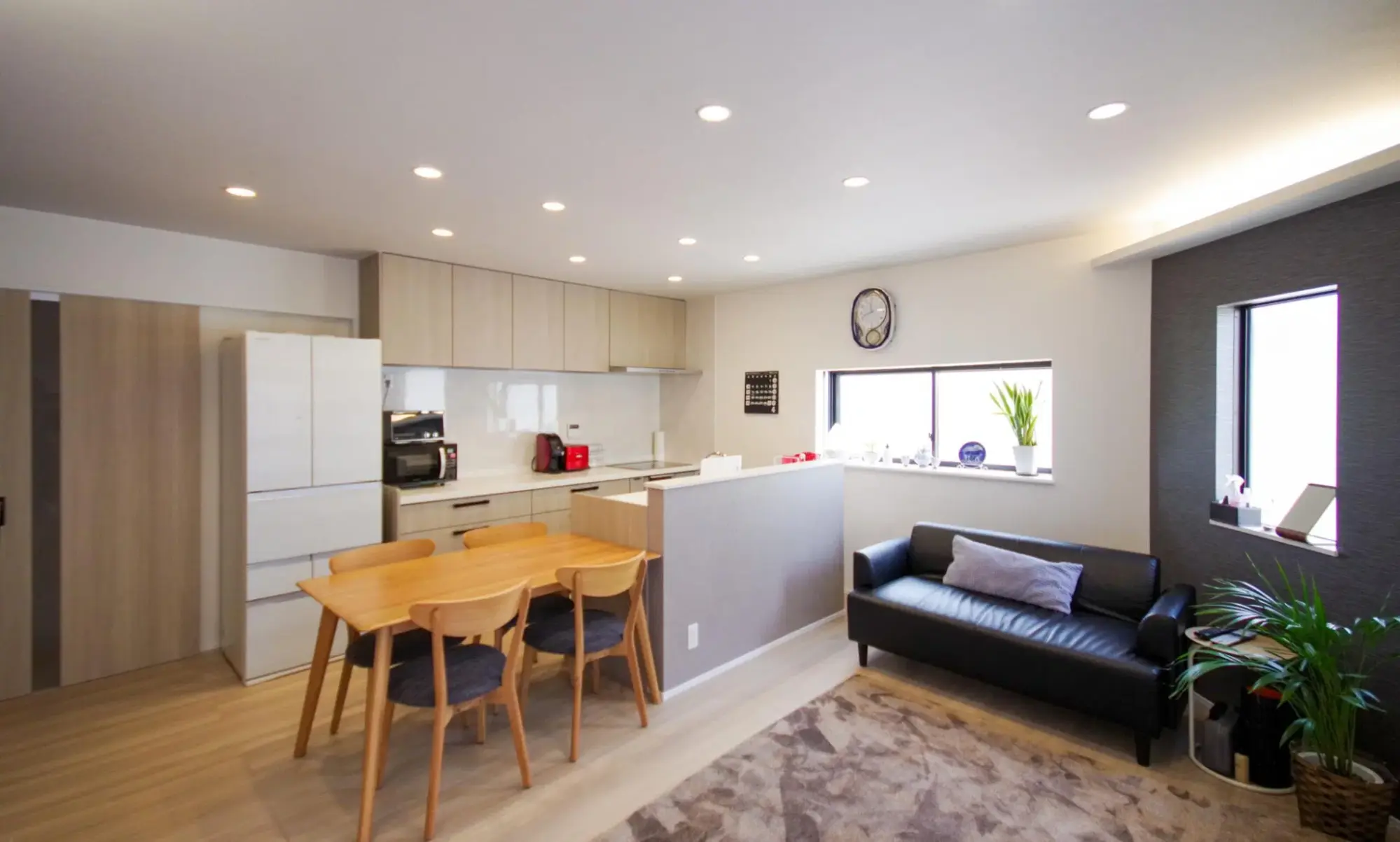 戸建てリノベーション後。木製ダイニングと黒いソファの奥に、グレー腰壁の2列型対面キッチンを配置。ダークグレーのアクセントクロスと間接照明が空間を彩るナチュラルなLDK。リ・ワークショップのリノベーション事例。