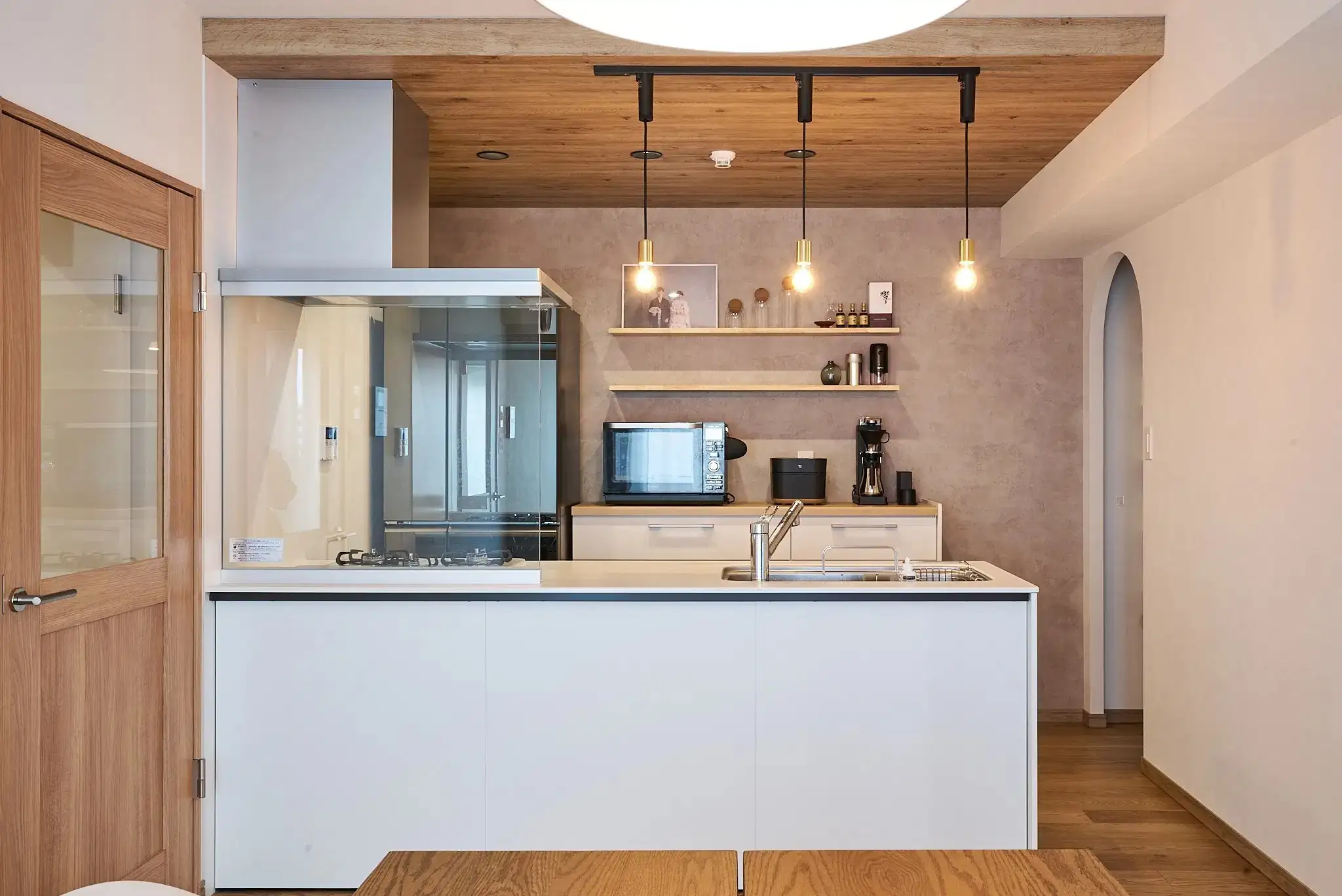 木目調の天井からペンダントライトが下がる、白を基調とした対面キッチン。背面には木製の飾り棚を設け、左手にはガラス入りの木製ドア、右手にはアーチ状の開口部を配置したマンションリノベーション内観。ANRI STYLE/リノベ図書館の施工事例。