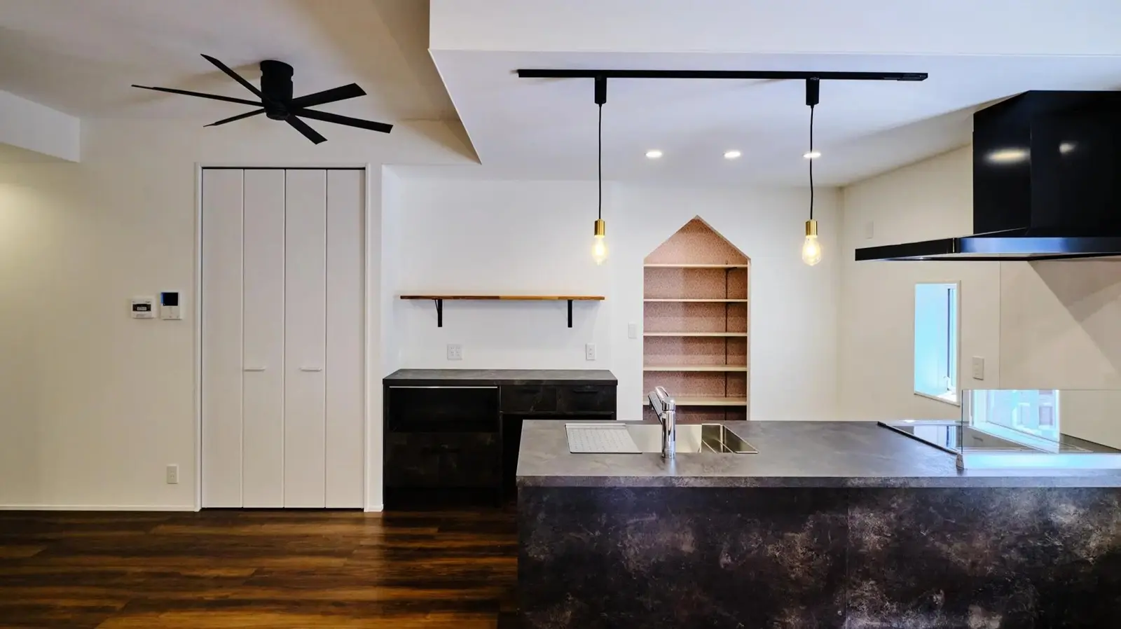 重厚感のあるダークグレーのグラフテクトキッチンを配置したLDK。背面のパントリー入口は三角垂れ壁のデザインになっており、黒いシーリングファンやペンダントライトが空間を引き締めている、大人シックな倉庫リノベーションの内観。