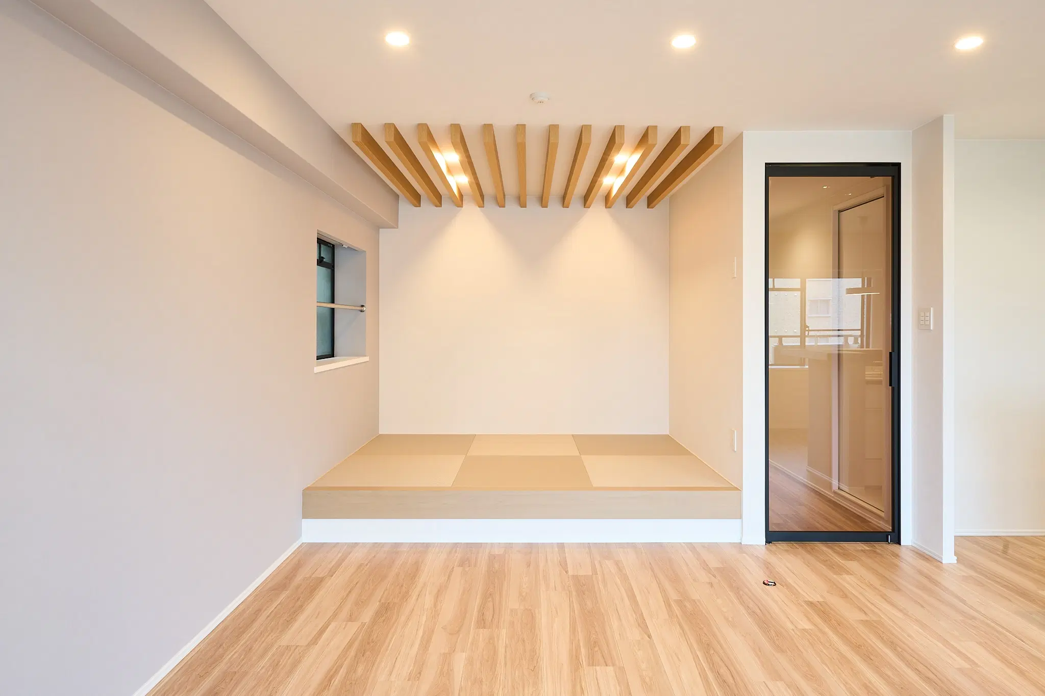 小上がりの畳スペースがあるLDKの写真。天井には木製のルーバーがあり、ダウンライトが設置されている。奥にはガラス扉があり、手前の床は明るい木目調。