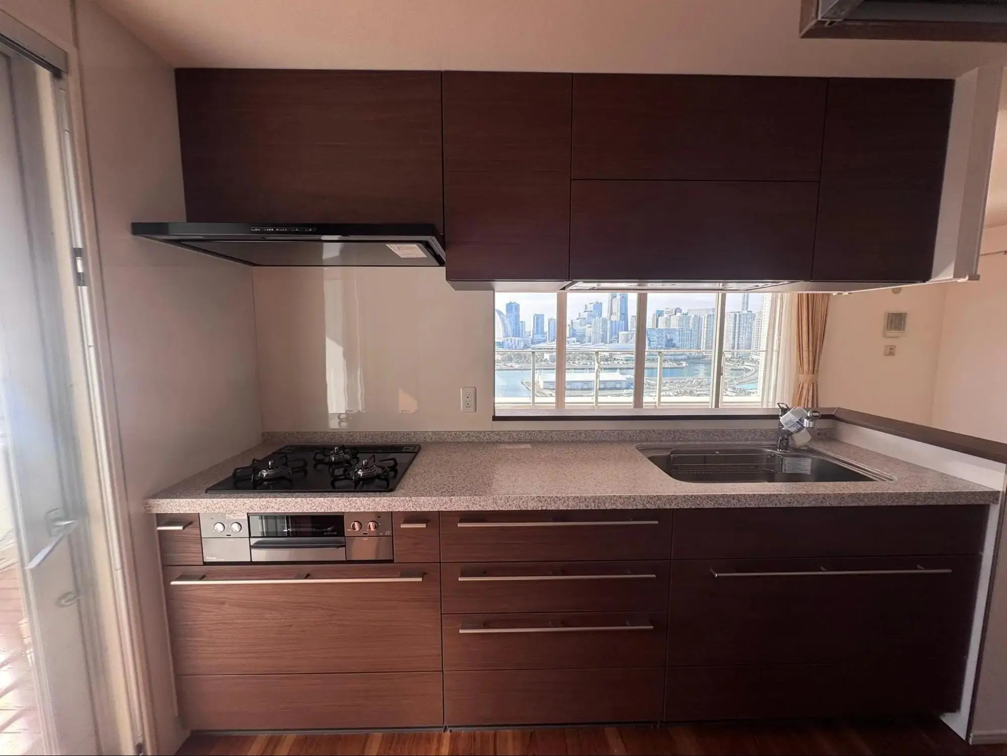 3口ガスコンロと広々としたシンクを備えた、石目調ワークトップのキッチン。作業スペースの正面にある窓からは、海とみなとみらいのビル群を一望できる。料理中も景色を楽しめるタワーマンションリノベーション。リフレネクスト横浜の施工事例。