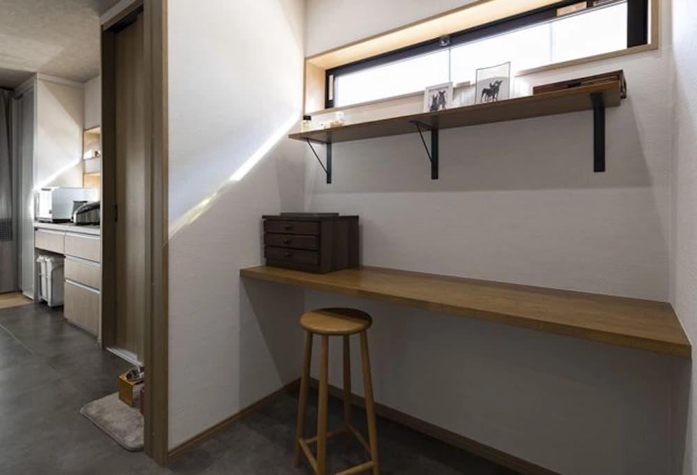 キッチンのすぐ横に設けられた、シンプルで機能的なカウンターヌック。壁面に備え付けられた長い木製カウンターの前には、背の高い丸い木製スツールが置かれている。カウンターの上部には、黒いアイアンのブラケットで支えられた2段の飾り棚があり、お気に入りの小物をディスプレイできる。さらにその上には横長の高窓が設置されており、外からの視線を遮りながらも安定した自然光を空間に取り込んでいる。床は落ち着いたダークグレーのタイル調で、キッチンへと続く開口部にはペット用の水飲みボウルが置かれており、家族とペットが共生する温かみのある暮らしを感じさせる空間。