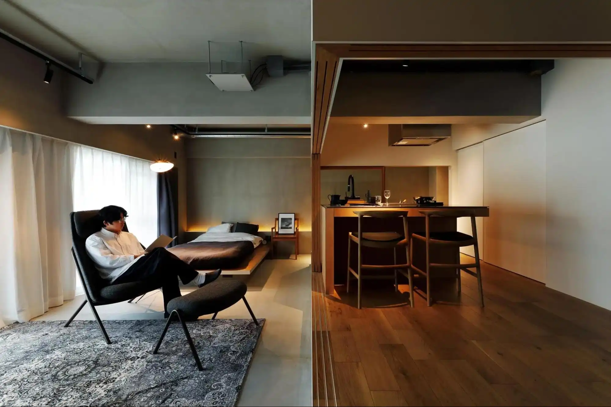 ワンルームマンションのLDKと寝室の写真。手前にラウンジチェアに座る男性、奥にベッド、右側に小上がりのキッチンカウンターが配置されている。