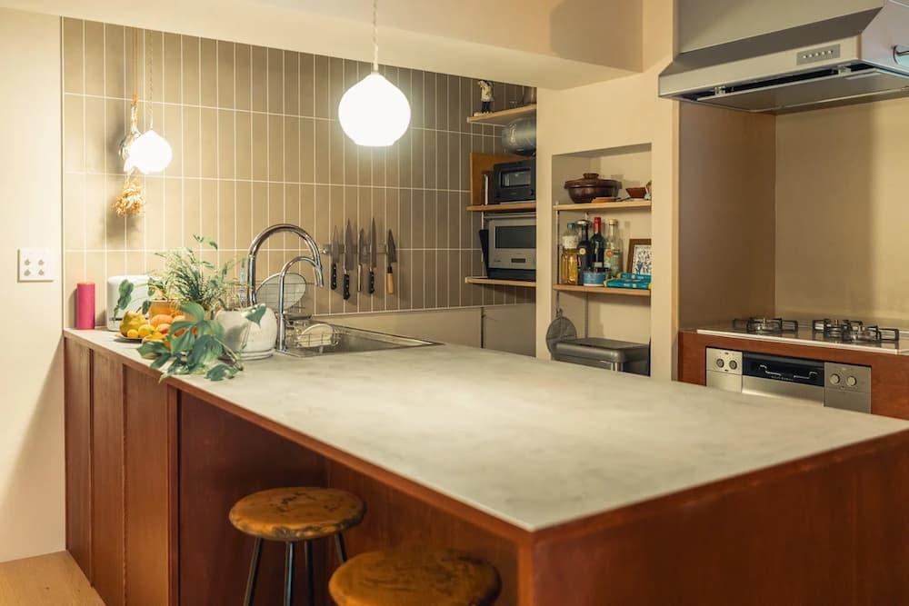 モルタル調のグレー天板と赤茶色の木製ベースを組み合わせた、こだわりの造作対面キッチン。壁面には落ち着いた色味の縦長タイルをあしらい、丸いペンダントライトやヴィンテージスツールがカフェのような温かみを演出している。ステンレスのレンジフードや壁面のナイフホルダーなど、機能美とインダストリアルな要素が調和した内観。