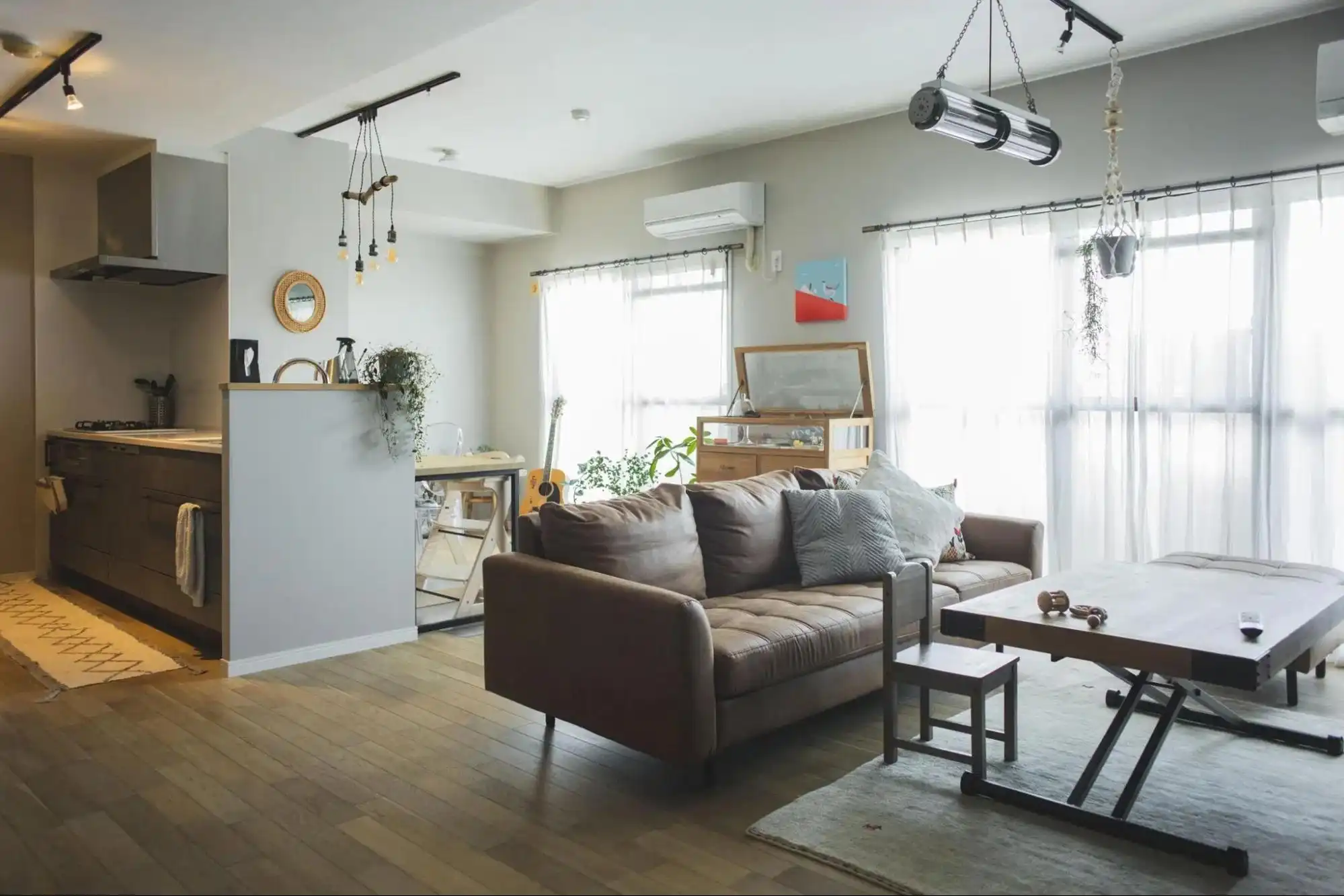 LDKの写真。茶色の革製ソファと木製テーブルが置かれ、奥には木製カウンターのキッチンが見える。壁は薄いグレーで床は木製。