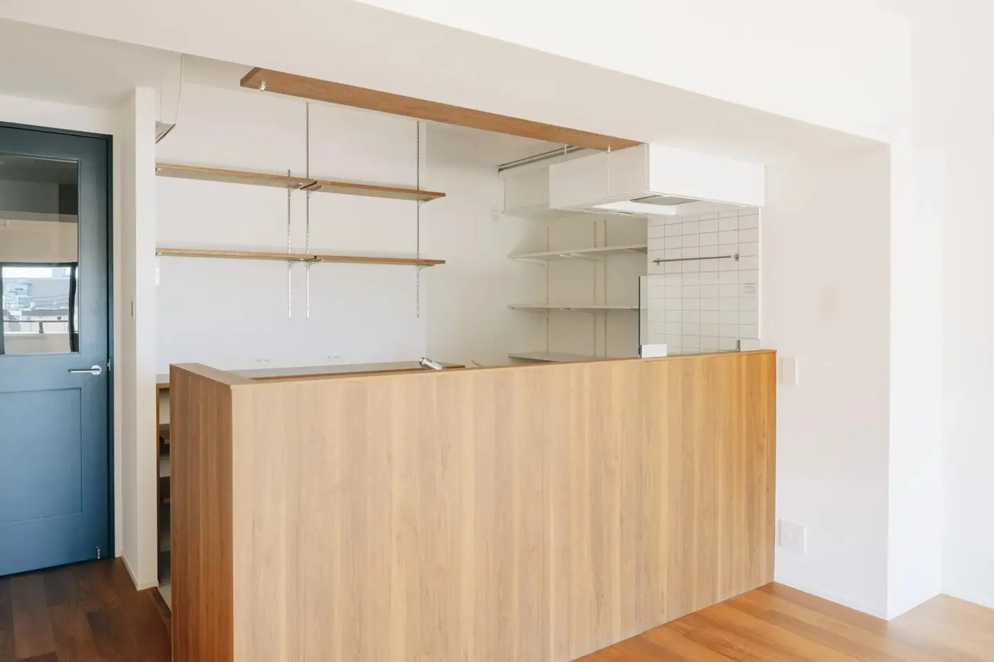 fujitacaリノベーションの事例。チーク材のフローリングと木目の対面キッチンが温かみを与えるLDK。背面には白いタイルとオープンシェルフを配置し、左手のブルーグレーのドアがアクセント。既存設備を活かしデザインを一新したマンションリノベ内観。