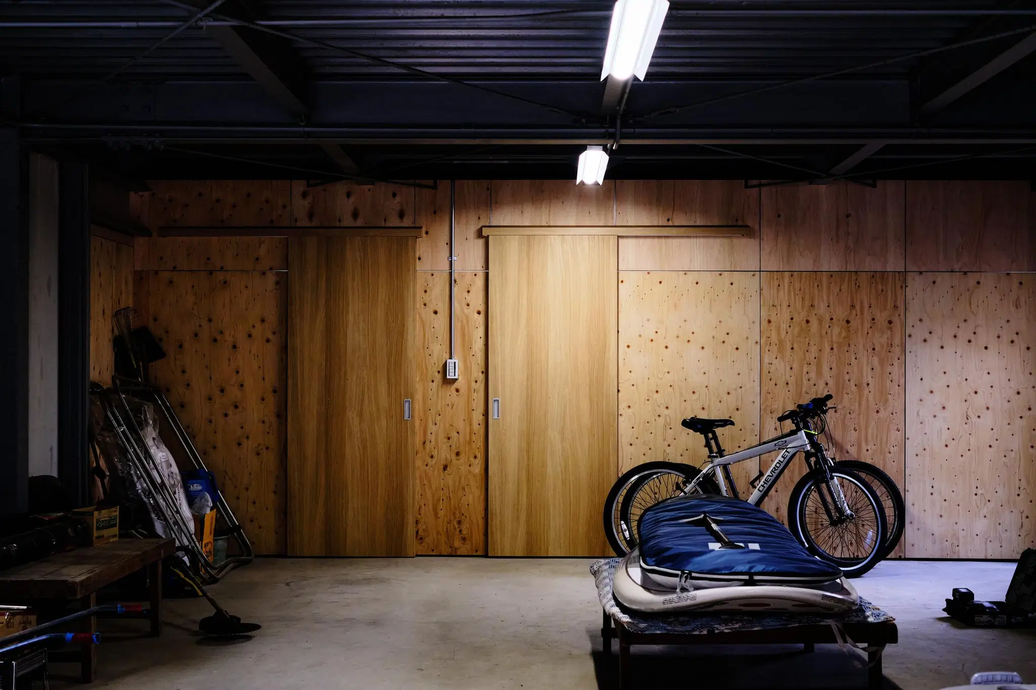 倉庫リノベーション後の1階インナーガレージ。コンクリート床と構造用合板の壁面、2枚の木製引き戸で構成。黒い鉄骨天井の下に自転車やアウトドア用品が並ぶインダストリアルな空間。株式会社松岡住研|リノベースのリノベーション事例。