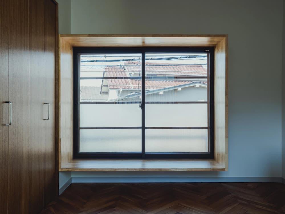 深い色味のヘリンボーン床と白い壁が美しいコントラストを描く室内。窓枠を囲うように明るい色合いの木材で厚みのあるフレームを造作し、腰掛けて外を眺められるベンチ型のウィンドウヌック。黒枠の引き違い窓には格子状の横桟と曇りガラスが組み合わされ、隣接する大容量の木製クローゼットとともに、素材の質感を大切にしたミニマルで和モダンな雰囲気のリノベーション内観。