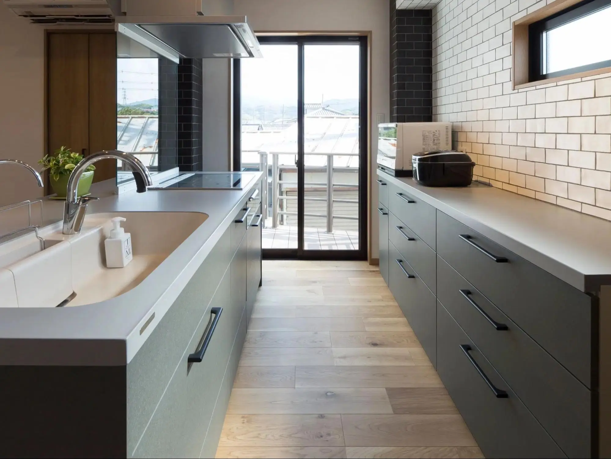Ⅱ型キッチン。マットグレーのキャビネットを中心に、右壁には白いサブウェイタイルを施工。奥の大きなガラス戸から自然光が差し込む、明るく落ち着いた空間。an cube（アン キューブ）のリノベーション事例。