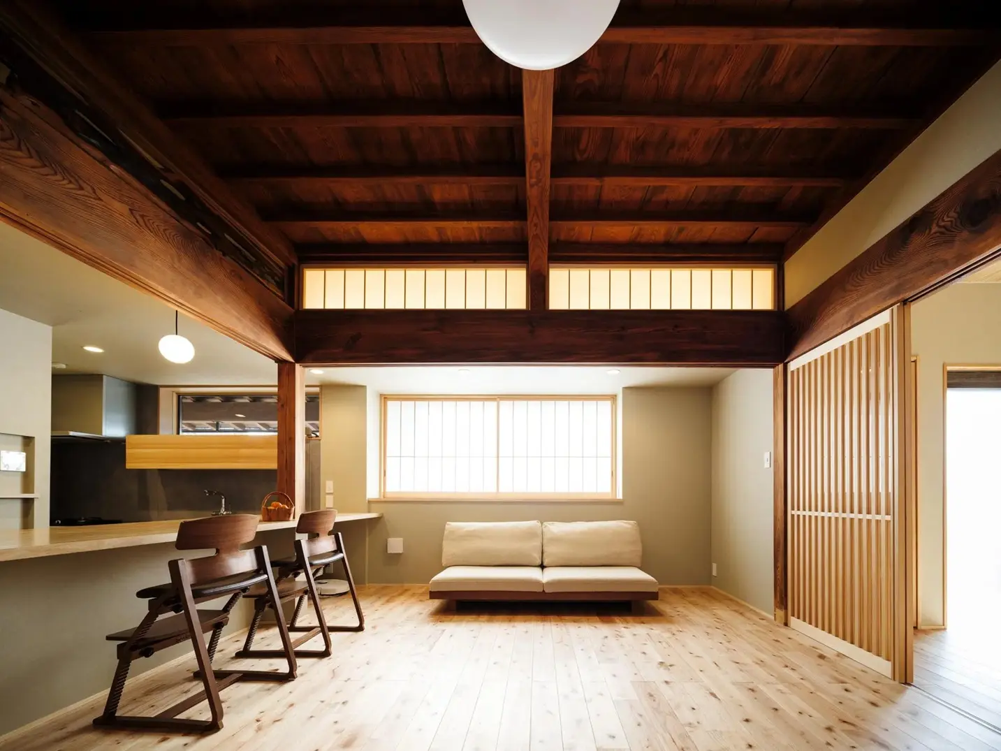 障子から柔らかな光が入るリビングスペース。天井近くの欄間窓や太い梁が古民家の歴史を感じさせる中、シンプルでモダンなベージュのローソファが置かれた落ち着いた空間。