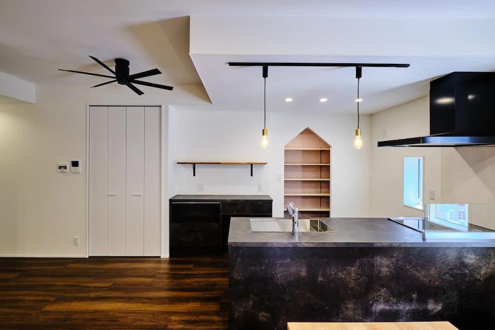 ダークグレーの石目調キッチンと黒いレンジフードが主役の、モダンでインダストリアルなLDKの内観。天井には黒のライティングレールとシーリングファン、真鍮ソケットの裸電球照明が設置され、無骨な雰囲気を強調している。白い壁と濃い木目の床で構成された空間の奥には、特徴的な家型の木製ニッチ棚があり、クールな中にユニークなアクセントを添えている。