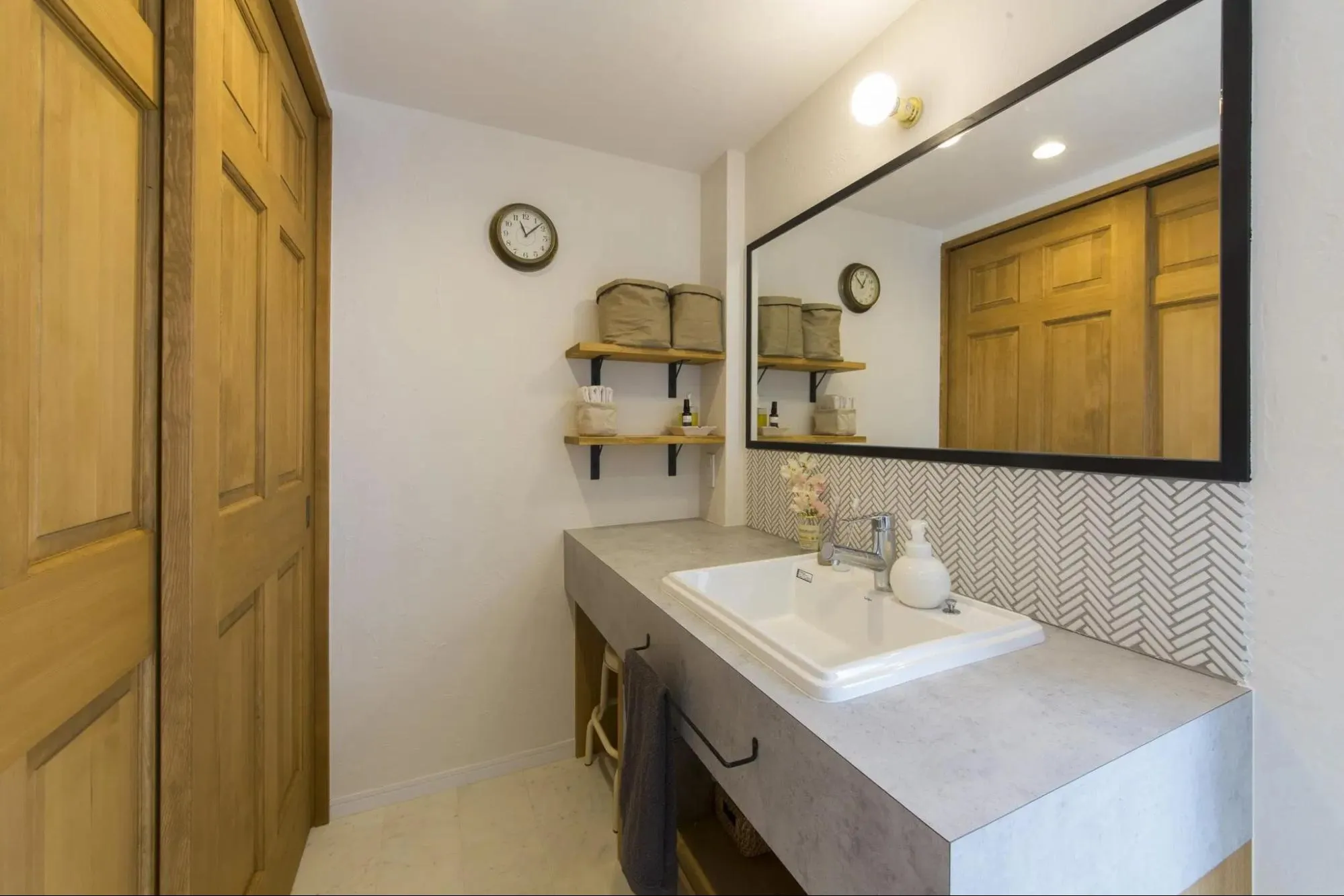 モルタル調のカウンターとヘリンボーン柄のタイル壁がスタイリッシュな造作洗面台。黒フレームの鏡や木製ドア、オープン棚を組み合わせ、シンプルで使い勝手の良い空間に仕上げたリノベーション。パパママハウスの施工事例。