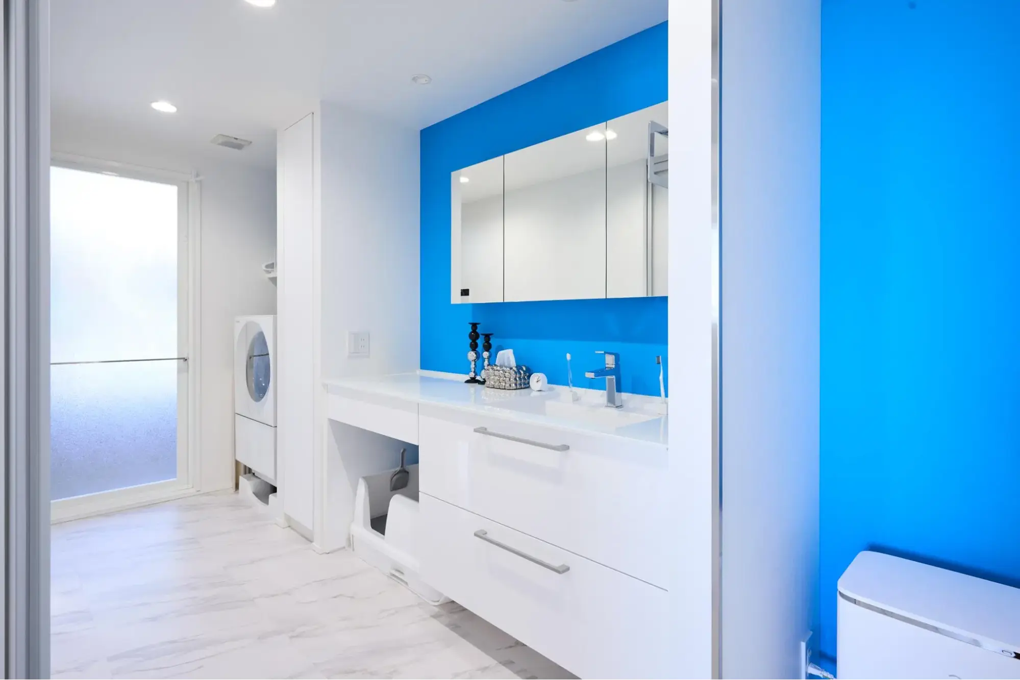 鮮やかなブルーのアクセントクロスが目を引く、白を基調としたホテルライクな洗面室。ご自身で商品を組み合わせたオリジナルの洗面台は、カウンター下部に愛猫のトイレをすっきりと配置できるスペースを確保している。猫との暮らしと美しいデザインを両立させた、ANRI STYLE/リノベ図書館のマンション施工事例。