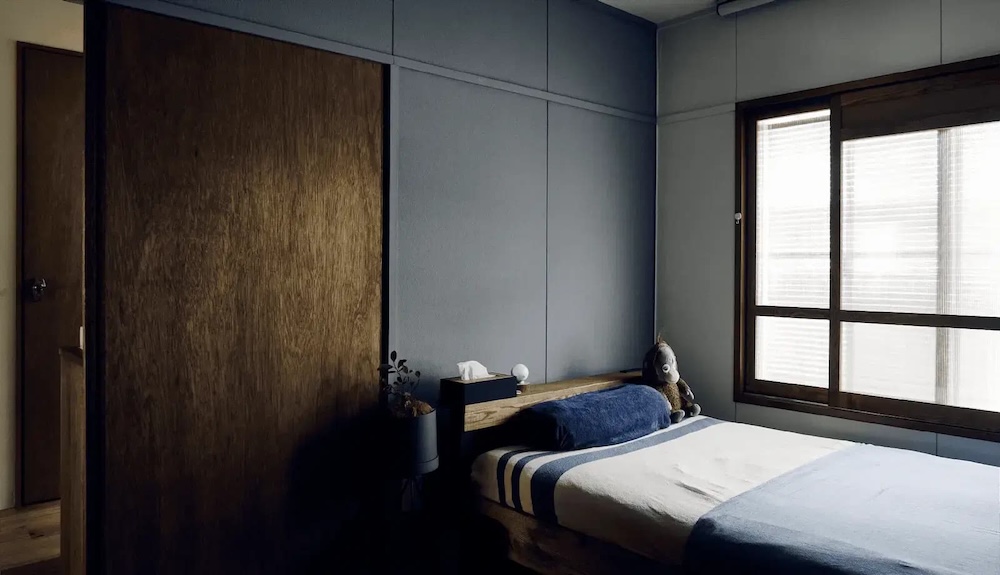 落ち着いたブルーグレーのアクセントウォールと、木目が美しいヴィンテージ風の大きな引き戸が絶妙にマッチした、インダストリアルテイストの寝室。木枠の窓にはブラインドを合わせ、ネイビーと白のベッドリネンでカラーを統一した、DIYやセルフリノベーションの参考にもなる、シンプルで男前なベッドルームインテリア実例。