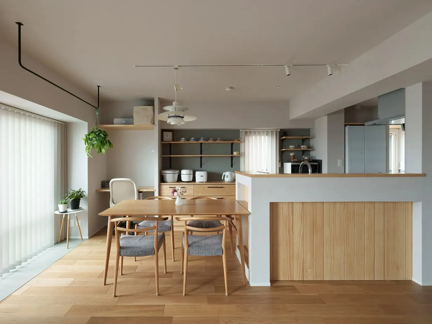 木目の腰壁が特徴的な対面キッチンと、グレーのチェアを合わせたダイニング。奥にはワークスペースや見せる収納棚を配置。段差のないフラットな床で家事動線を改善した、北欧ナチュラルなマンションリノベ内観。インテリックス空間設計の施工事例