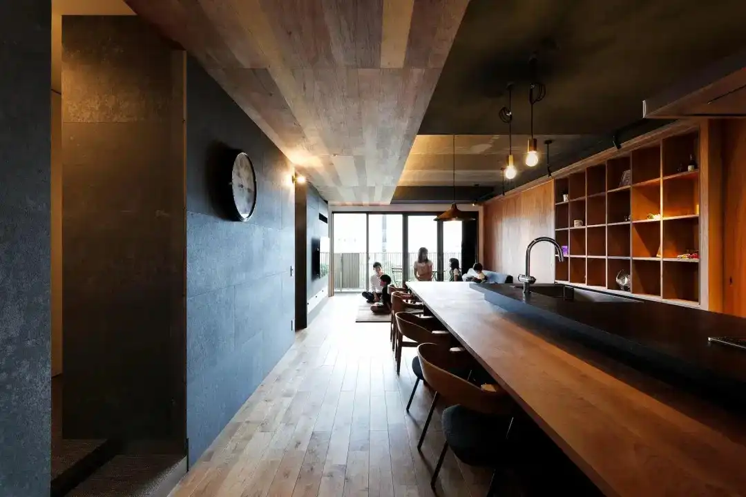 LDKの写真。長い木のカウンターバーがあり、床も木製。左の壁はダークグレーで、天井の一部も板張り。奥の窓際に家族が座っている。