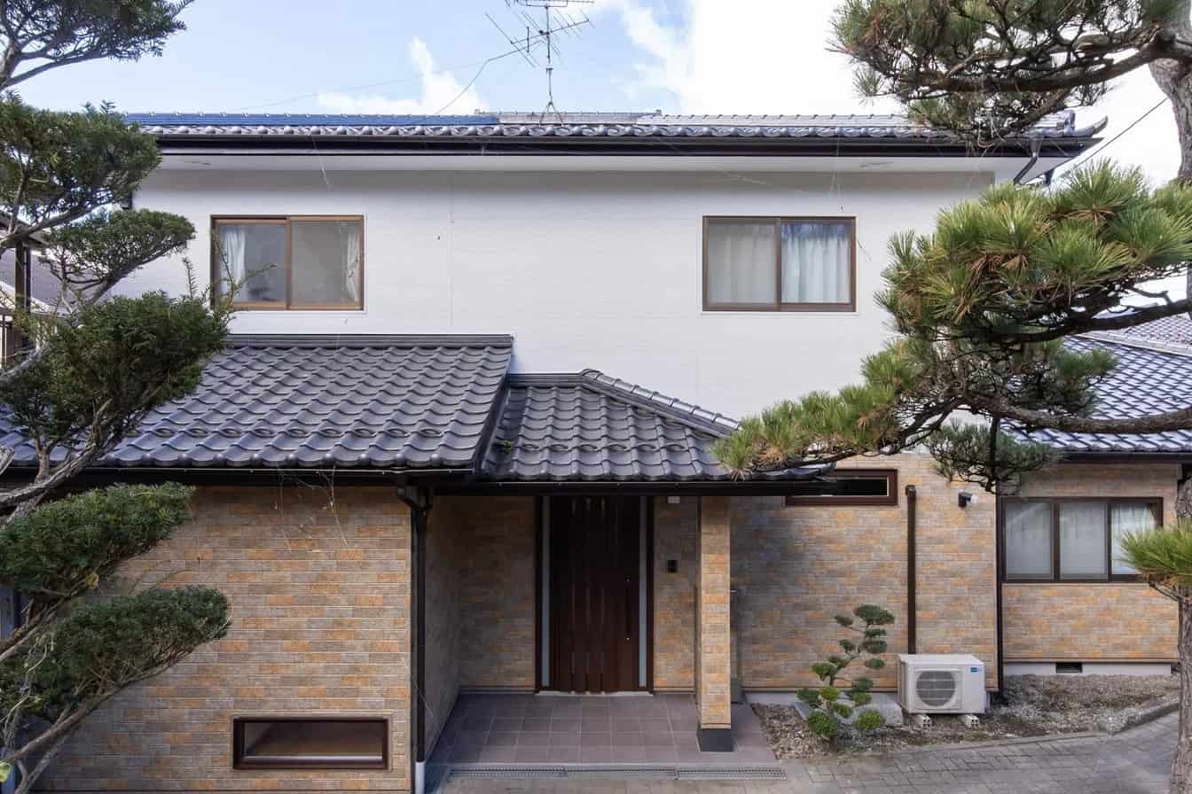 瓦屋根の二階建てで、レンガ調の外壁と白塗りの壁を組み合わせた現代的な日本家屋の外観。木製の玄関扉や木枠の窓があり、剪定された松や砂利の庭が和モダンな雰囲気を演出している。