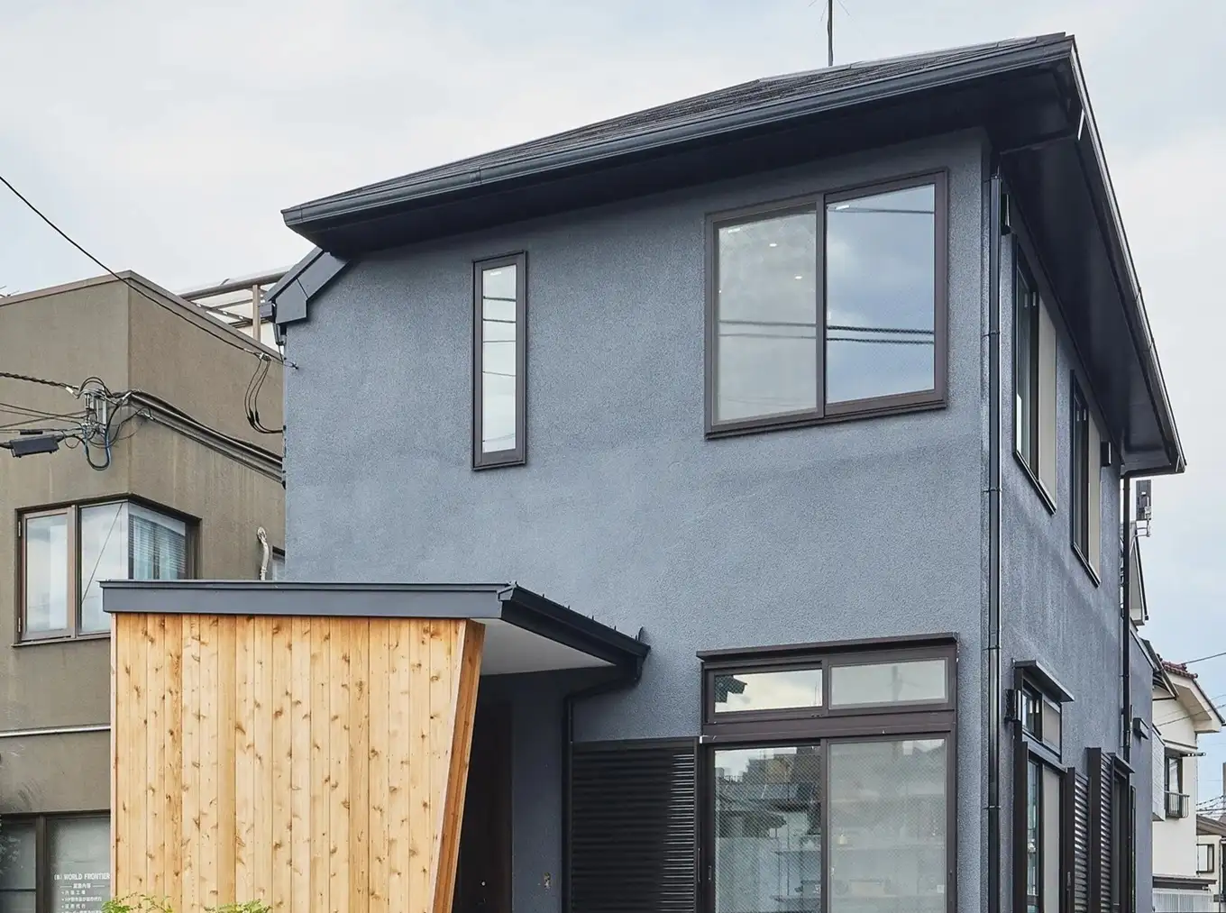 落ち着いたブルーグレーの吹き付け外壁に、斜めにカットされた木目調の玄関ポーチがアクセントの外観。既存の窓の形状を活かしつつ、シックなカラーリングと天然木の質感でスタイリッシュに仕上げた、築18年の戸建てフルリノベーション事例。