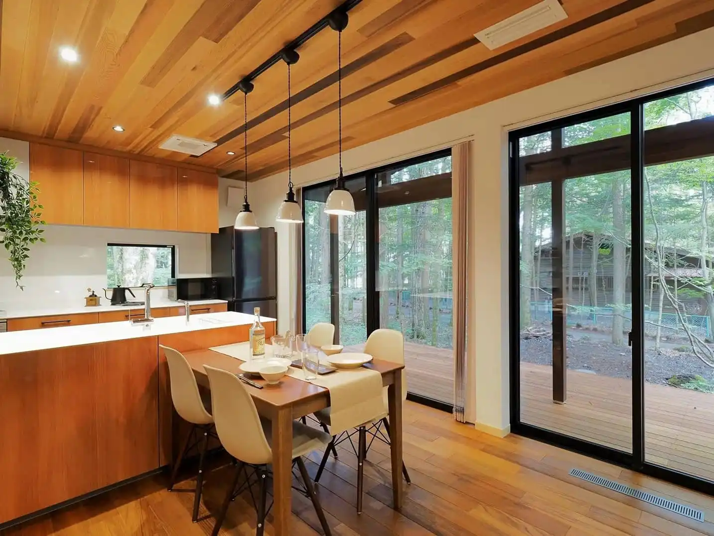 天井まである大きな掃き出し窓から、美しい森の緑とウッドデッキを望む開放的なダイニングキッチン。板張りの勾配天井に黒いライティングレールとペンダントライトが映え、木の温もりに包まれた平屋リノベーション後の内観。