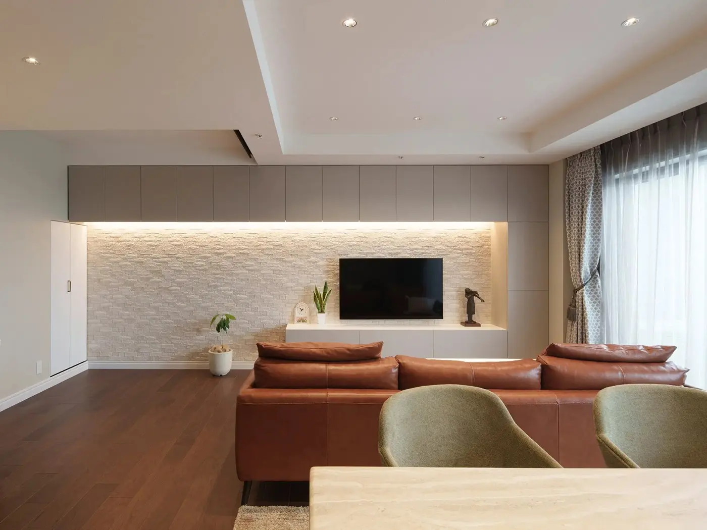 間接照明が照らすエコカラットの壁と、上下に設けた白い壁面収納が洗練された印象を与えるリビング。壁掛けテレビやブラウンのレザーソファを配置し、耐震性も考慮したホテルライクな空間。インテリックス空間設計の施工事例。