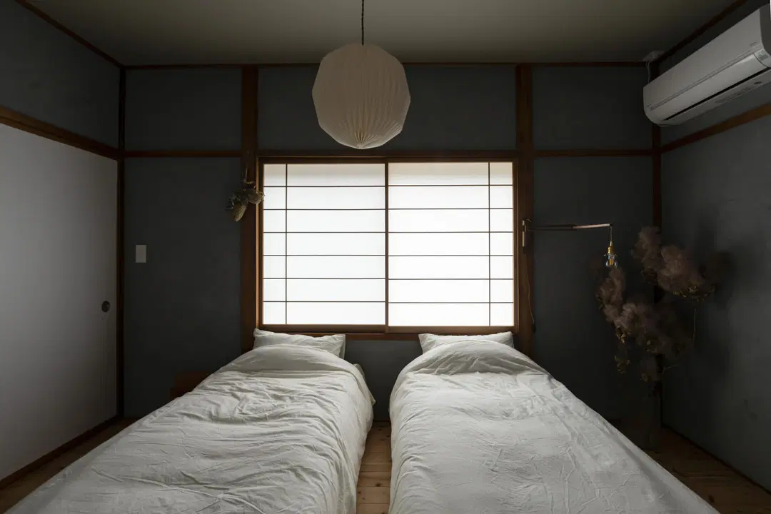 グレーの塗り壁と障子、無垢材の床を組み合わせた和モダンスタイル。ツインベッドと丸い和紙のペンダントライトが特徴的。