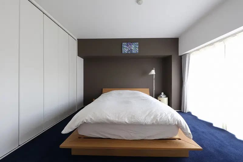 鮮やかなネイビーブルーのカーペットと、奥の壁面のダークグレーのアクセントクロスが都会的でクールな対比を生む、インダストリアルモダンな寝室。壁一面の白いクローゼットですっきりと見せつつ、木製のローベッド（プラットフォームベッド）と白いスタンドライトが清潔感と温かみを添える、ミニマルで洗練されたマンションリノベーションのインテリア実例。