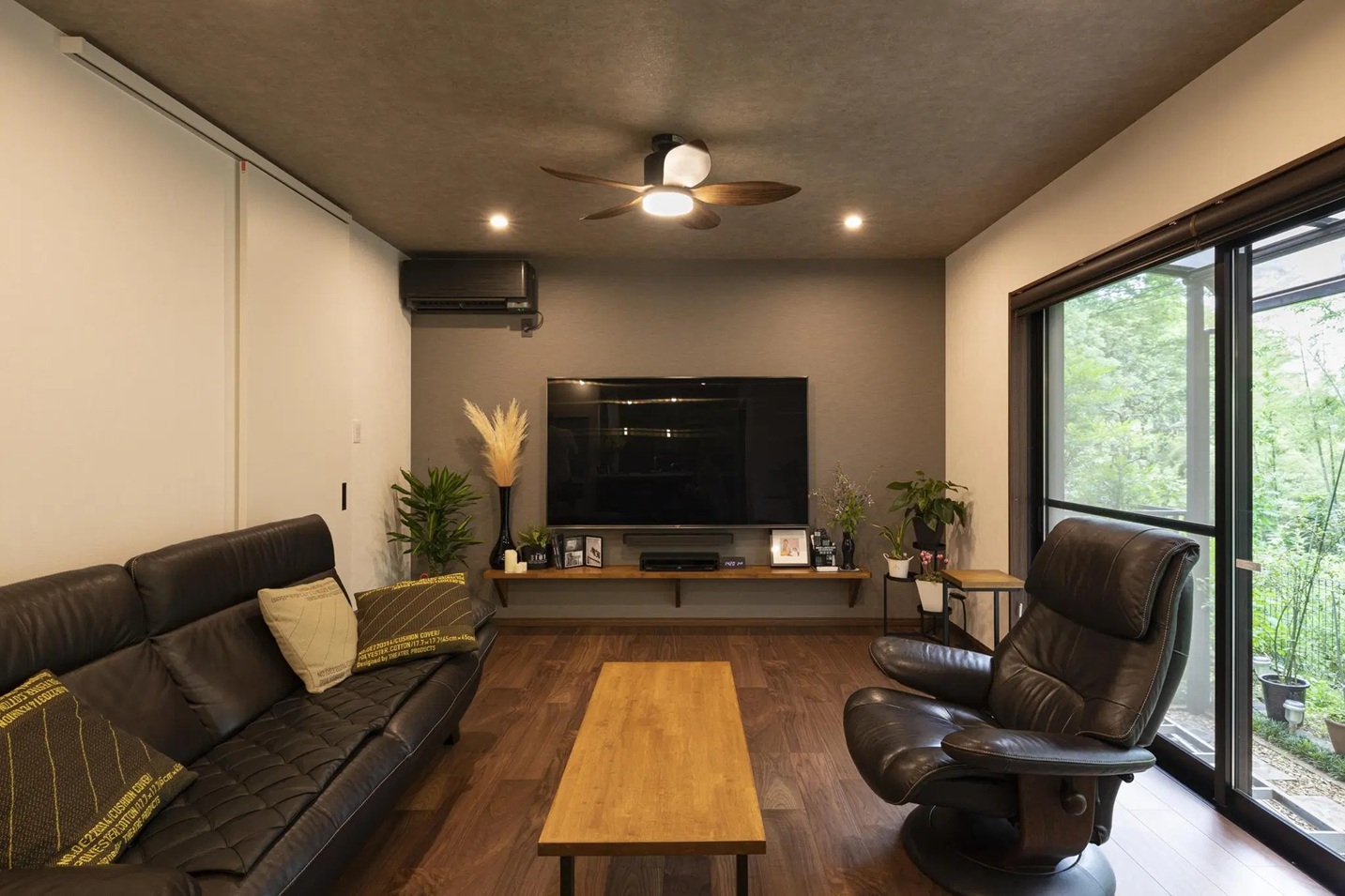 グレーのクロスで統一された壁・天井と、黒いレザーソファが重厚感を演出するリビング。木製のシーリングファンやテレビボードが温かみを添え、窓外の緑と調和したヴィンテージテイストの空間。an cubeの施工事例