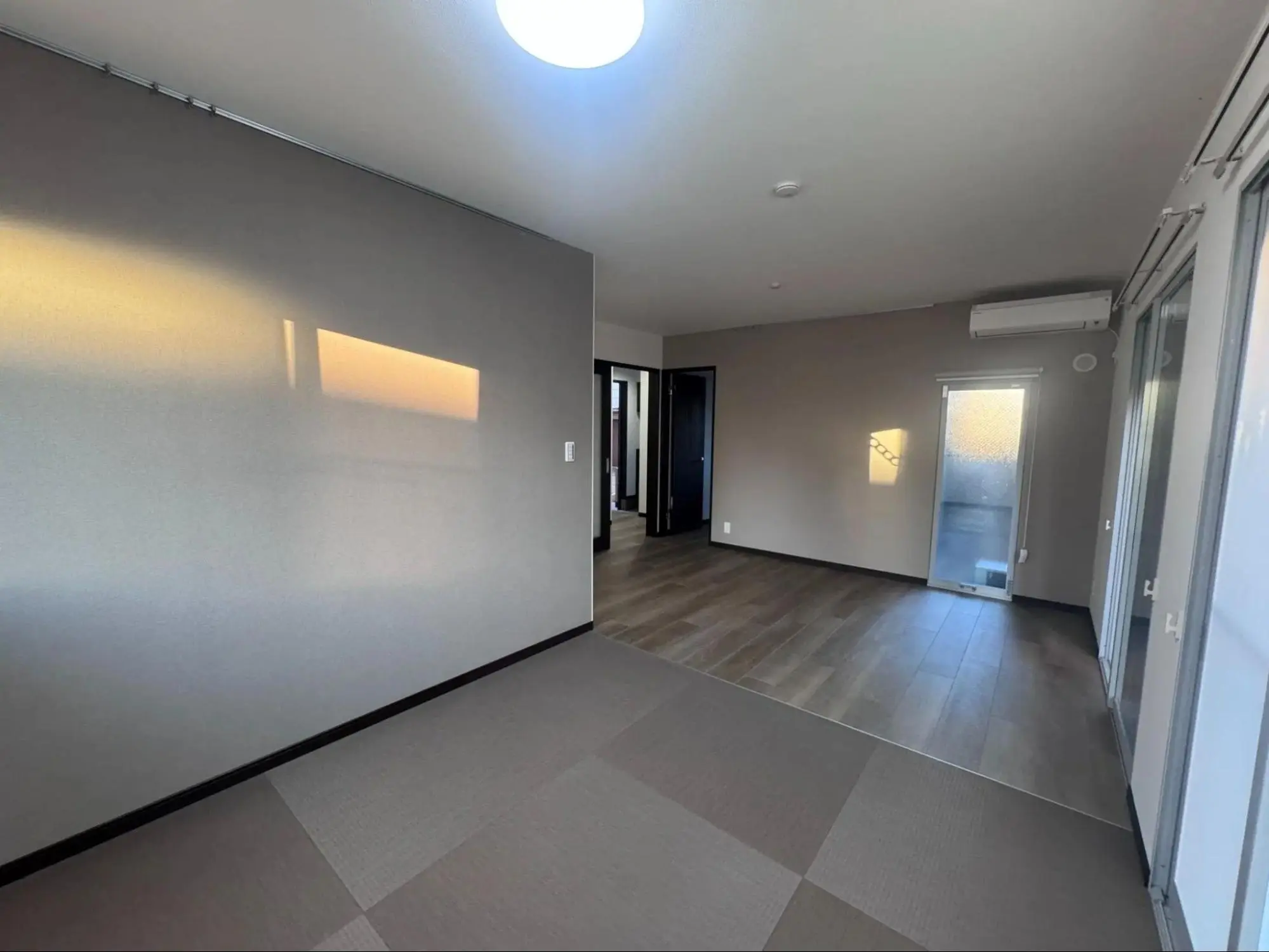 リビングルームの写真。壁はグレーで、床は木目調と市松模様のグレーの畳調の2種類。右手に窓が見える。