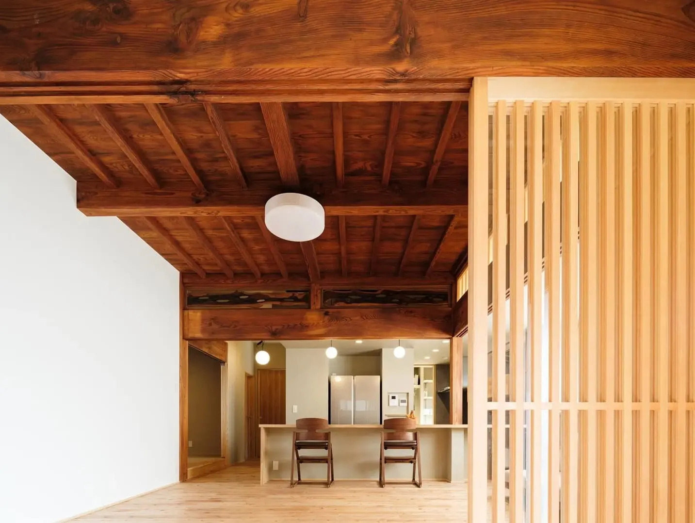 既存の太い梁と味わい深い板張り天井を活かした古民家の内装。空間を緩やかに仕切る縦格子の木製スクリーンと、白くモダンなシーリングライトが設置された、奥行きを感じる空間。