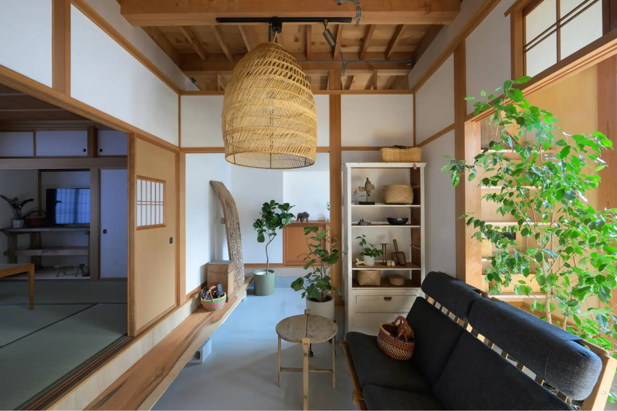戸建てフルリノベーション後。広い土間空間に黒いソファと白い棚を配置したセカンドリビング。左手には自然木の上がり框と和室が続く。木天井の大きな編み込み照明と多数の観葉植物が特徴的な和モダン空間。EUGUE RENOVATIONのリノベーション事例。