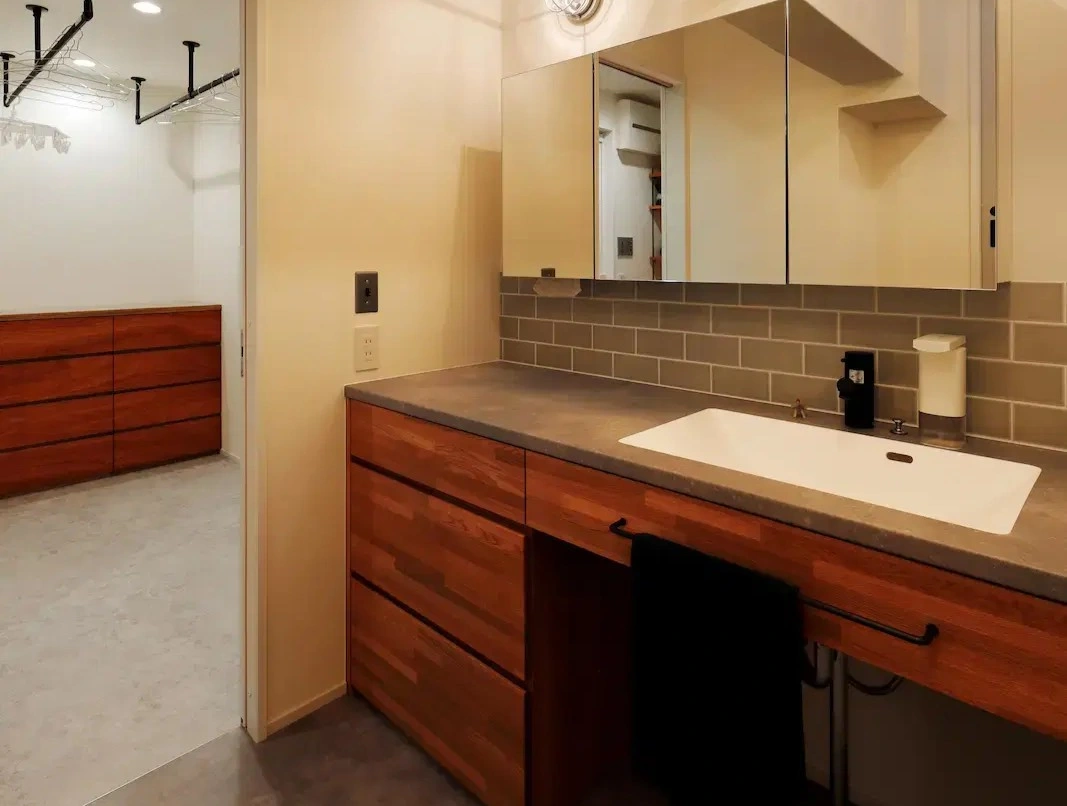 グレーの長方形タイルとモルタル調の天板、温かみのある木製キャビネットを組み合わせた造作洗面台。左手にはハンガーパイプを備えたファミリークローゼットが隣接し、広さと家事動線を確保したサニタリー空間。ひかり工務店の施工事例。